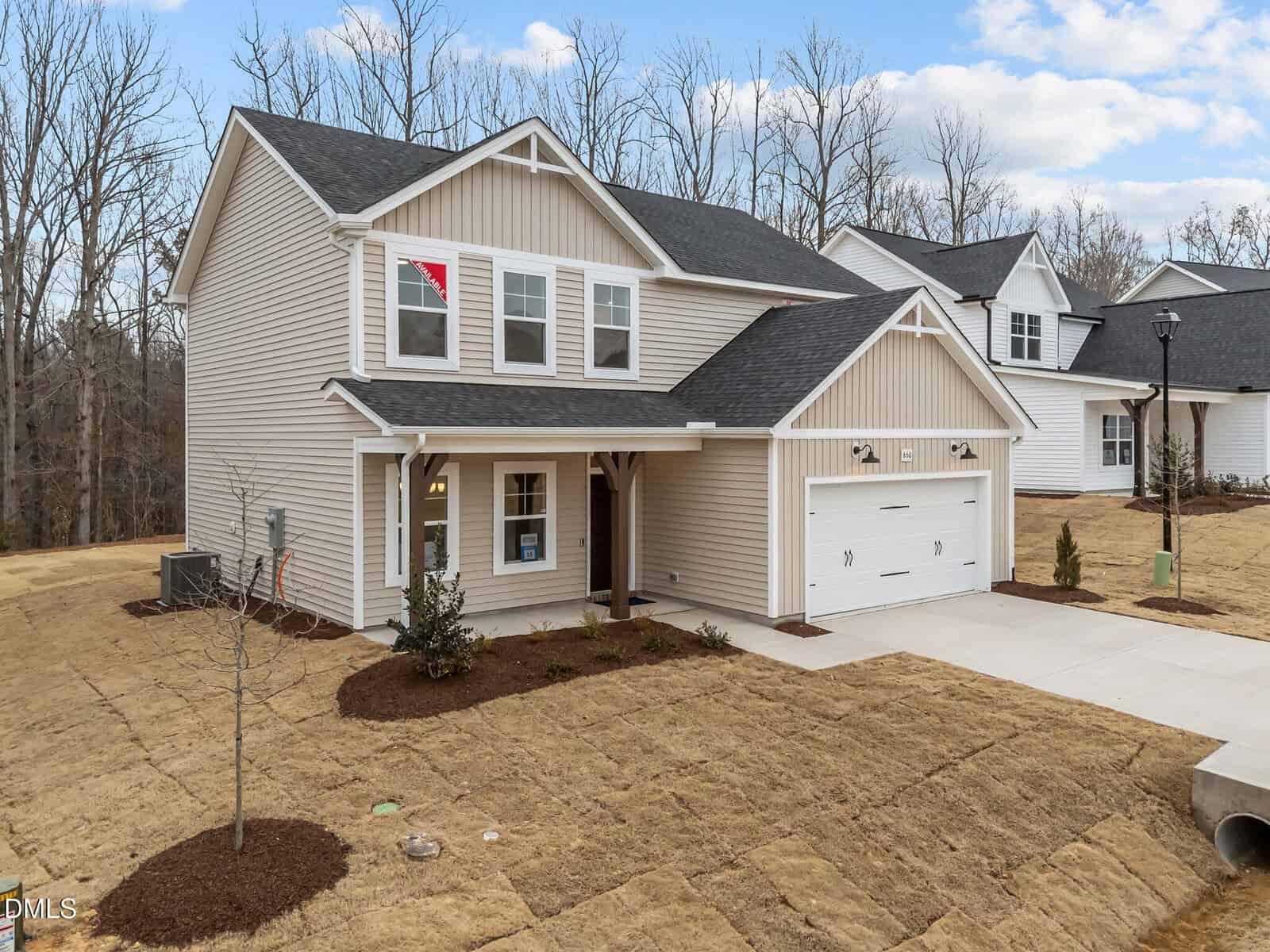 A two-story beige house with white trim, a covered front porch, a two-car garage, and a driveway. The yard is newly landscaped with mulch, young plants, and brown grass. Leafless trees are visible in the background.