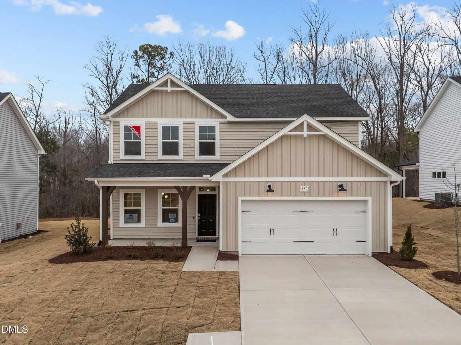 Two-story beige house with white trim, a covered front porch, and a two-car garage. The lawn is brown and there are a few small shrubs. Trees without leaves are visible in the background under a partly cloudy sky.