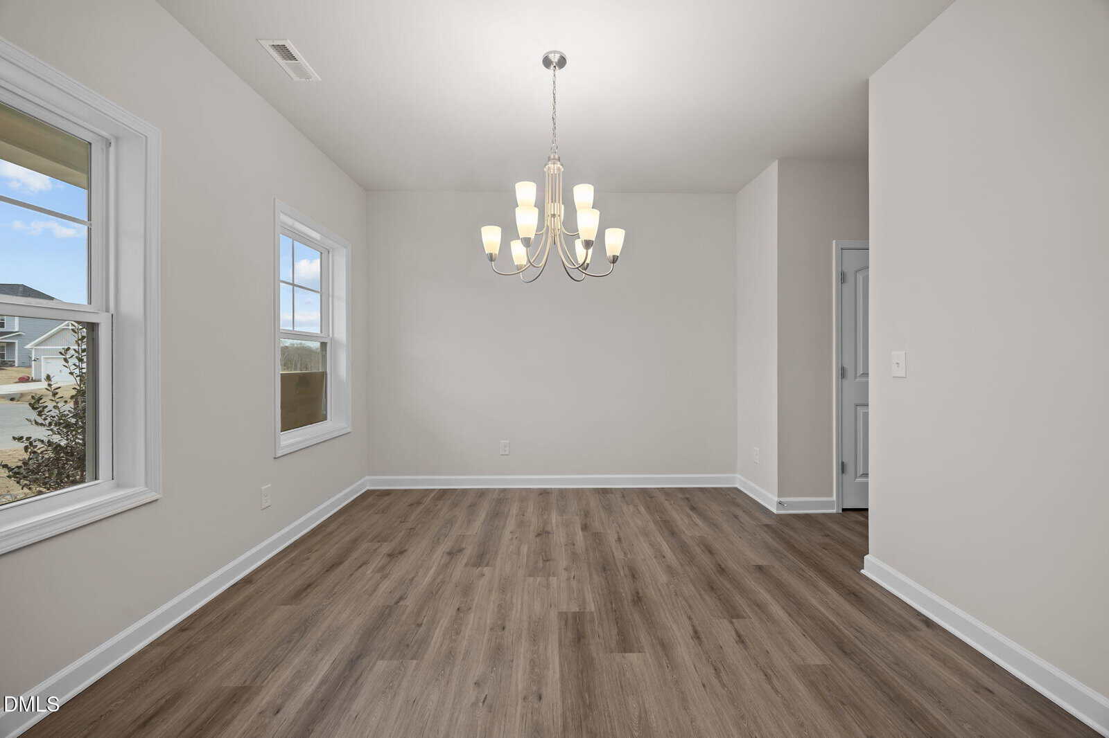 A dining room with light gray walls, wood-look vinyl flooring, two windows, a white baseboard, and a modern chandelier hanging from the ceiling. A doorway leads to another room.