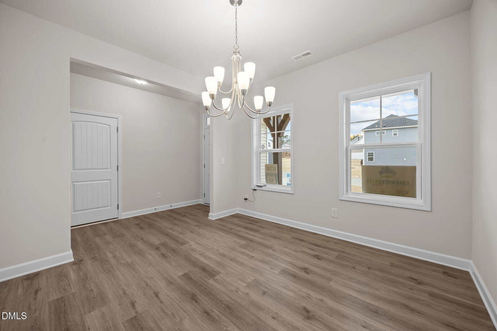 A bright, empty room with light gray walls, wood flooring, a white chandelier, two windows, and a white door. Natural light streams in, highlighting the clean, modern space.