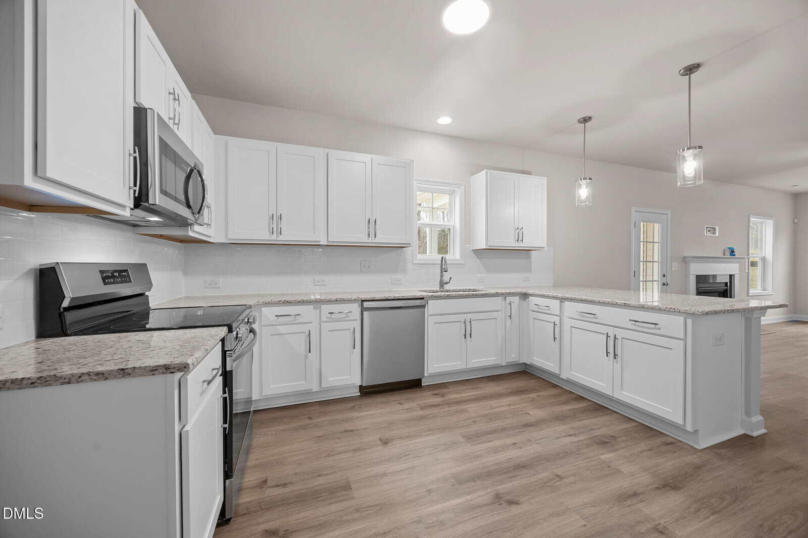 Modern kitchen with white cabinets, stainless steel appliances, granite countertops, and wood flooring. Pendant lights hang above a large peninsula, and a fireplace is visible in the open living area beyond.