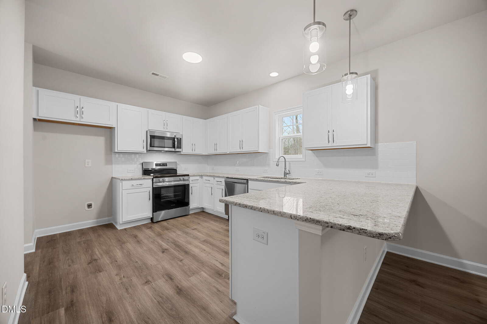Modern kitchen with white cabinets, stainless steel appliances, granite countertops, subway tile backsplash, pendant lights, and wood-look flooring. A large peninsula counter provides seating and workspace. Neutral walls and bright lighting.