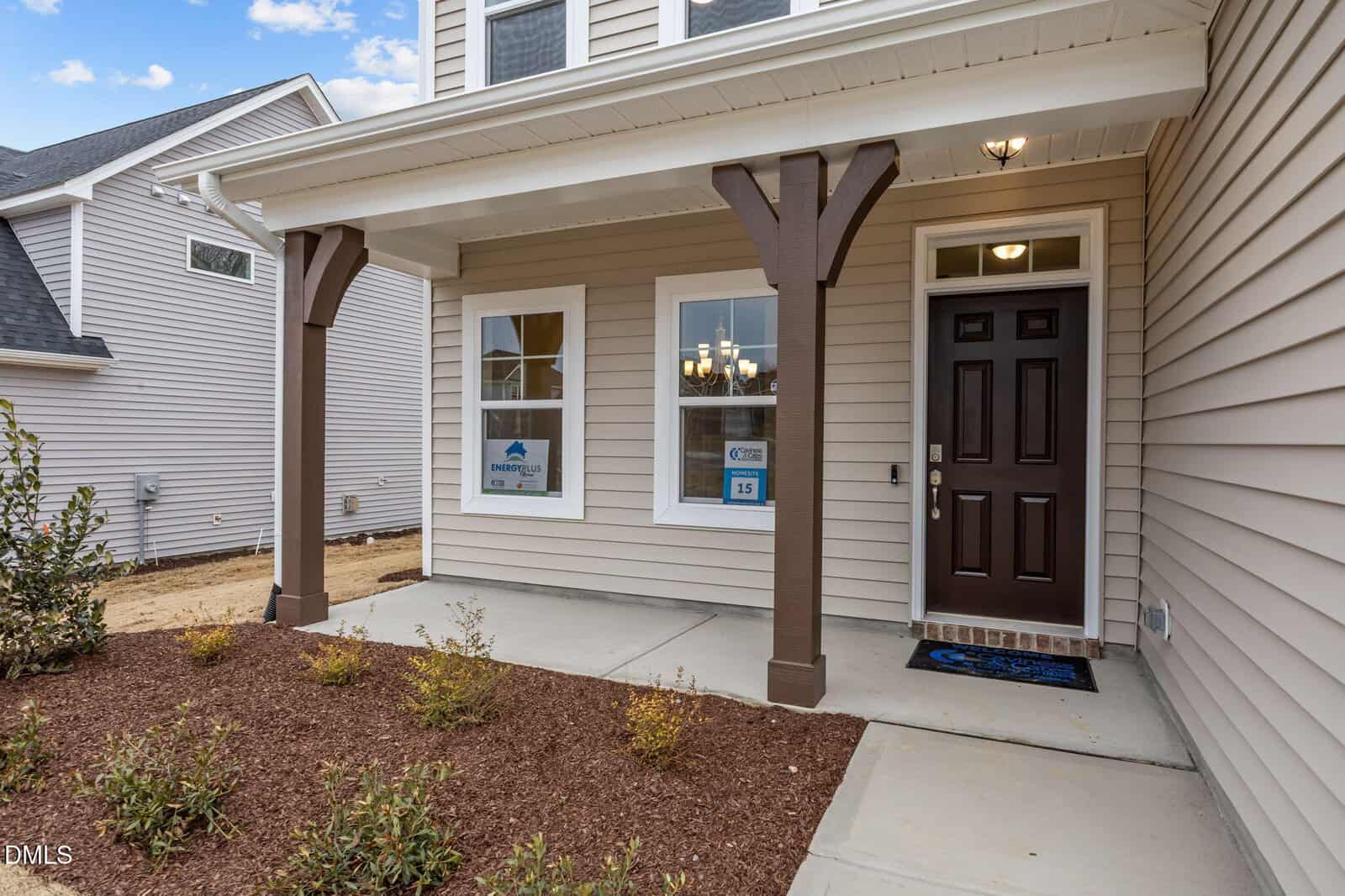 A beige single-family house features a covered front porch with two brown wooden posts, a black front door, two windows, mulch, small shrubs, and a blue welcome mat. Neighboring houses are partially visible.