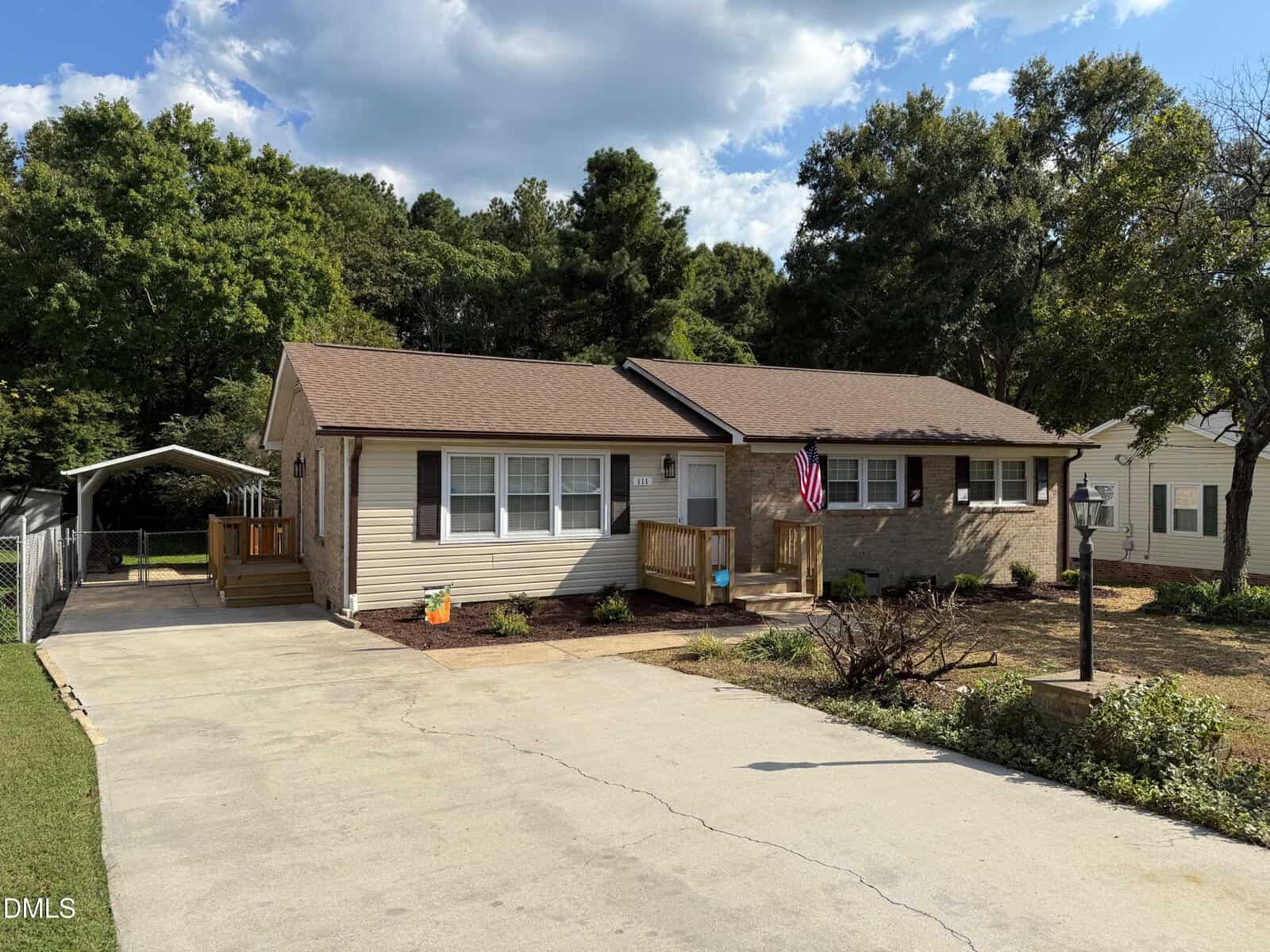 Charming single-story house at 111 Ward Avenue, Oxford, with beige siding, brown roof, concrete driveway, small front porch displaying an American flag, and a carport on the left. Trees and a partly cloudy sky complete the scene.