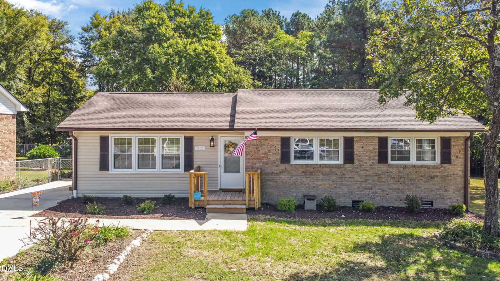 A single-story house with tan siding and brick exterior, dark brown shutters, and a brown roof. The front porch has a small wooden railing, two steps, and an American flag. There is a concrete walkway in front.