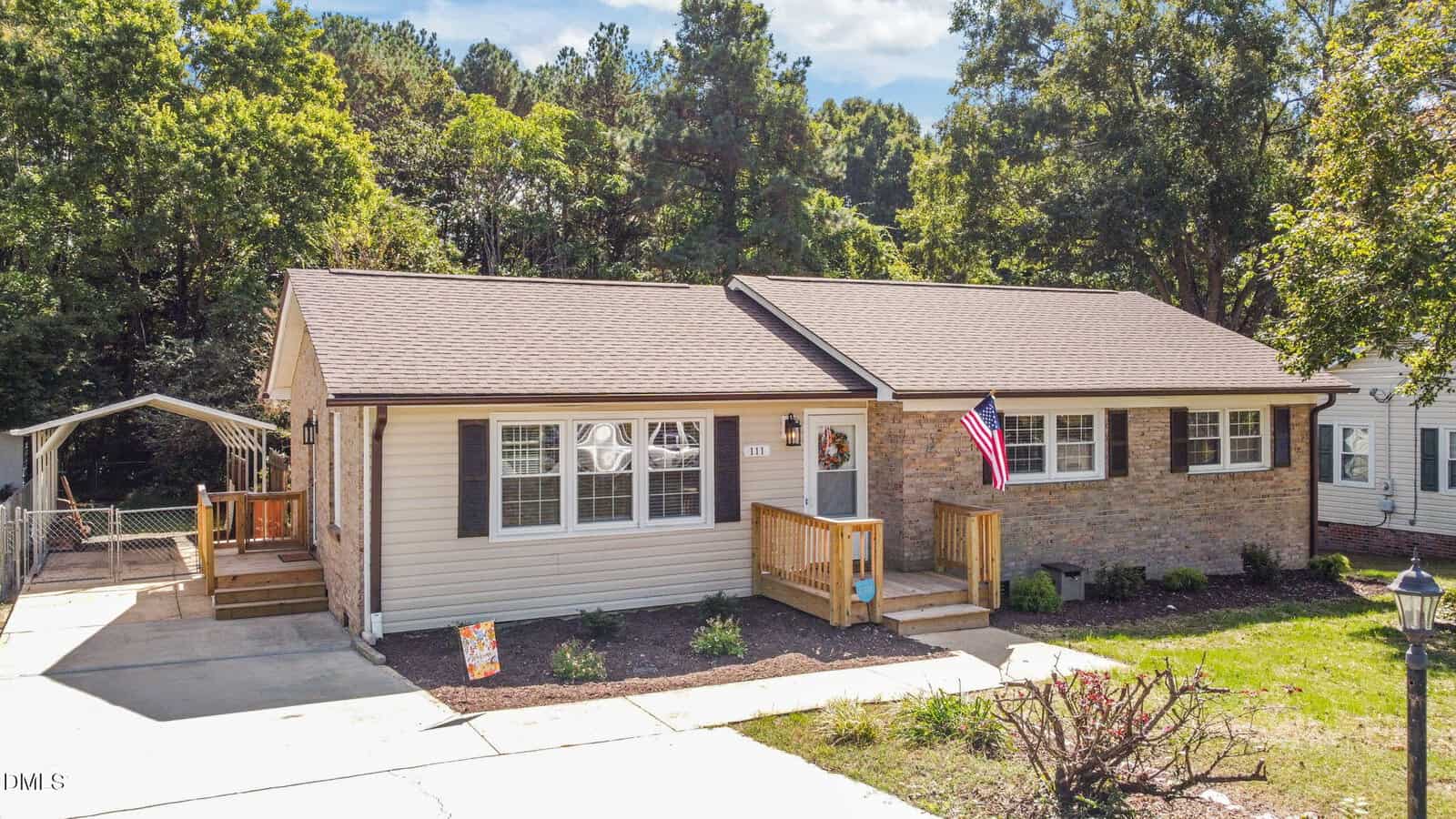 Charming single-story house at 111 Ward Avenue, Oxford, with light-colored siding and brick accents, a front porch with steps, an American flag, a paved driveway, surrounding trees, and a small shed or carport to the left.