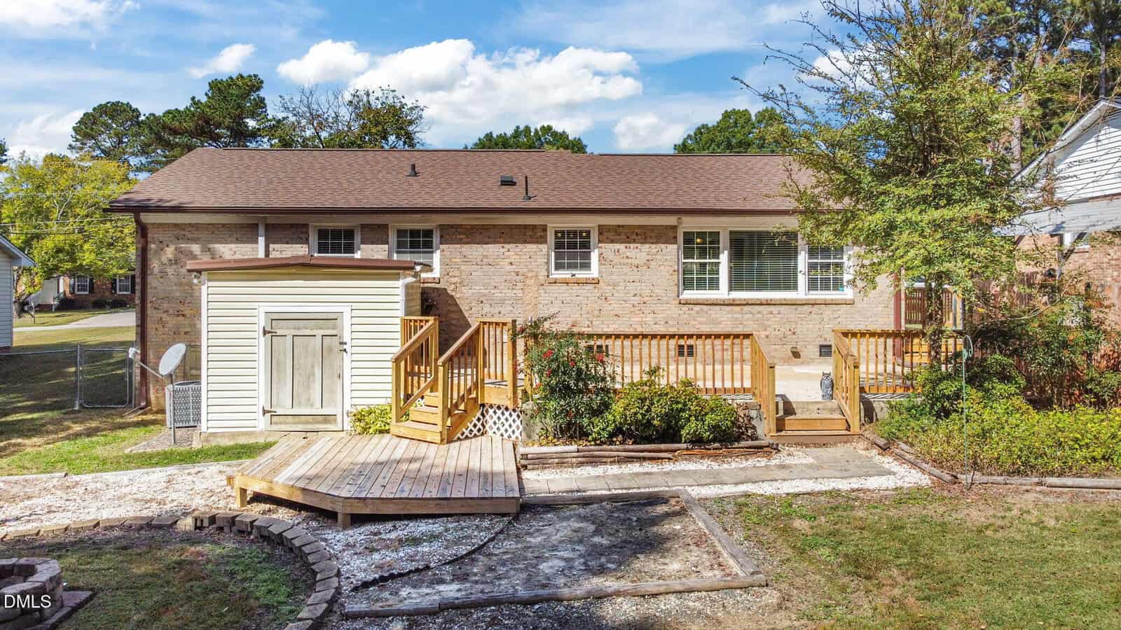 A single-story brick house with a brown roof, wooden deck, and small shed in the backyard. The yard includes landscaping, a paved path, and mature trees. The sky is partly cloudy.