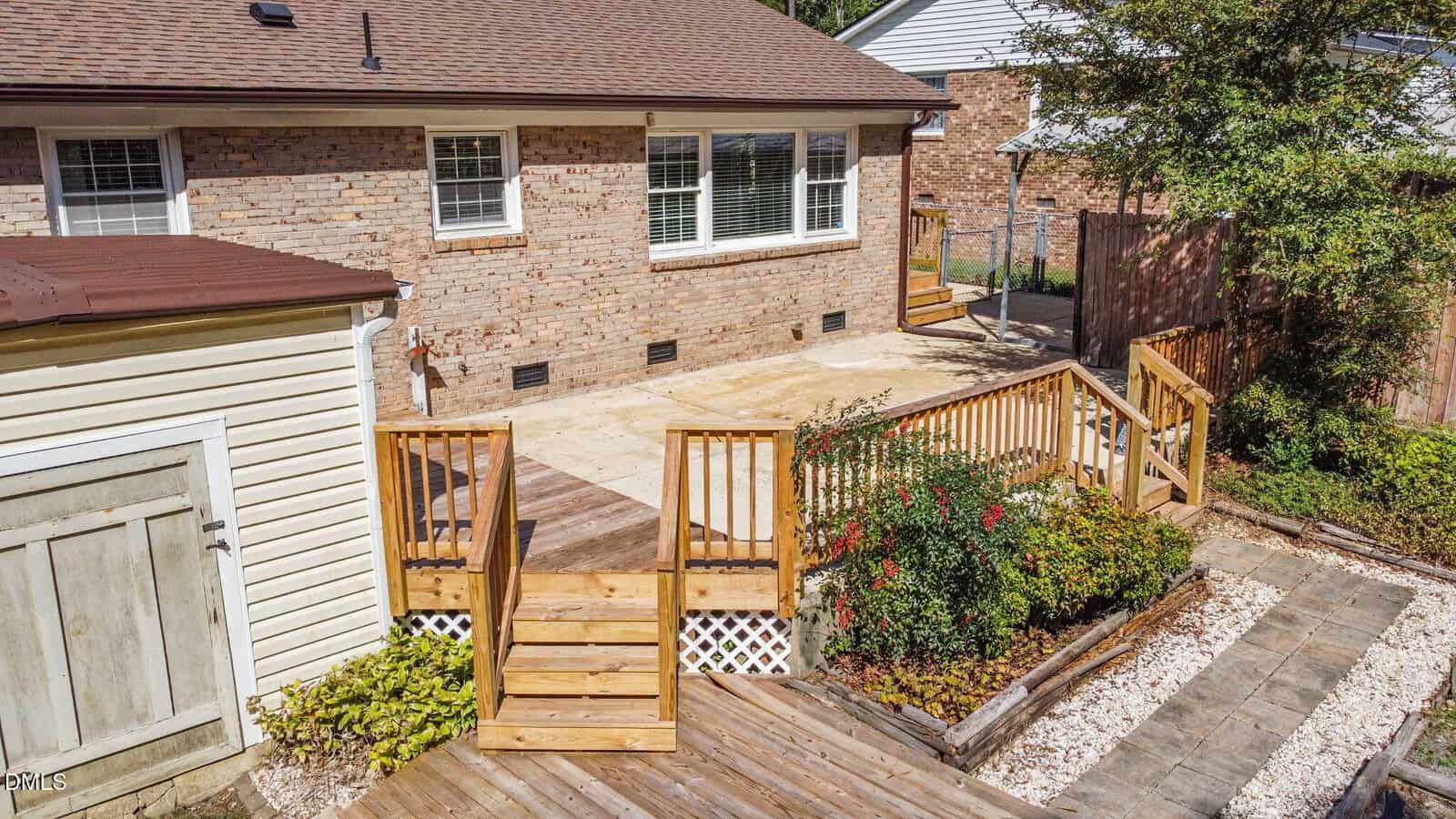 A backyard with a wooden deck and stairs leading to a concrete patio. The patio is adjacent to a brick house with three windows. There are garden beds with shrubs and a shed on the left side.