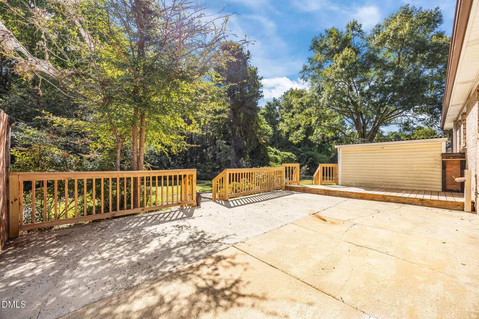 A concrete patio with wooden railings and steps leads to a small raised deck beside a beige outbuilding, surrounded by green trees under a sunny blue sky.