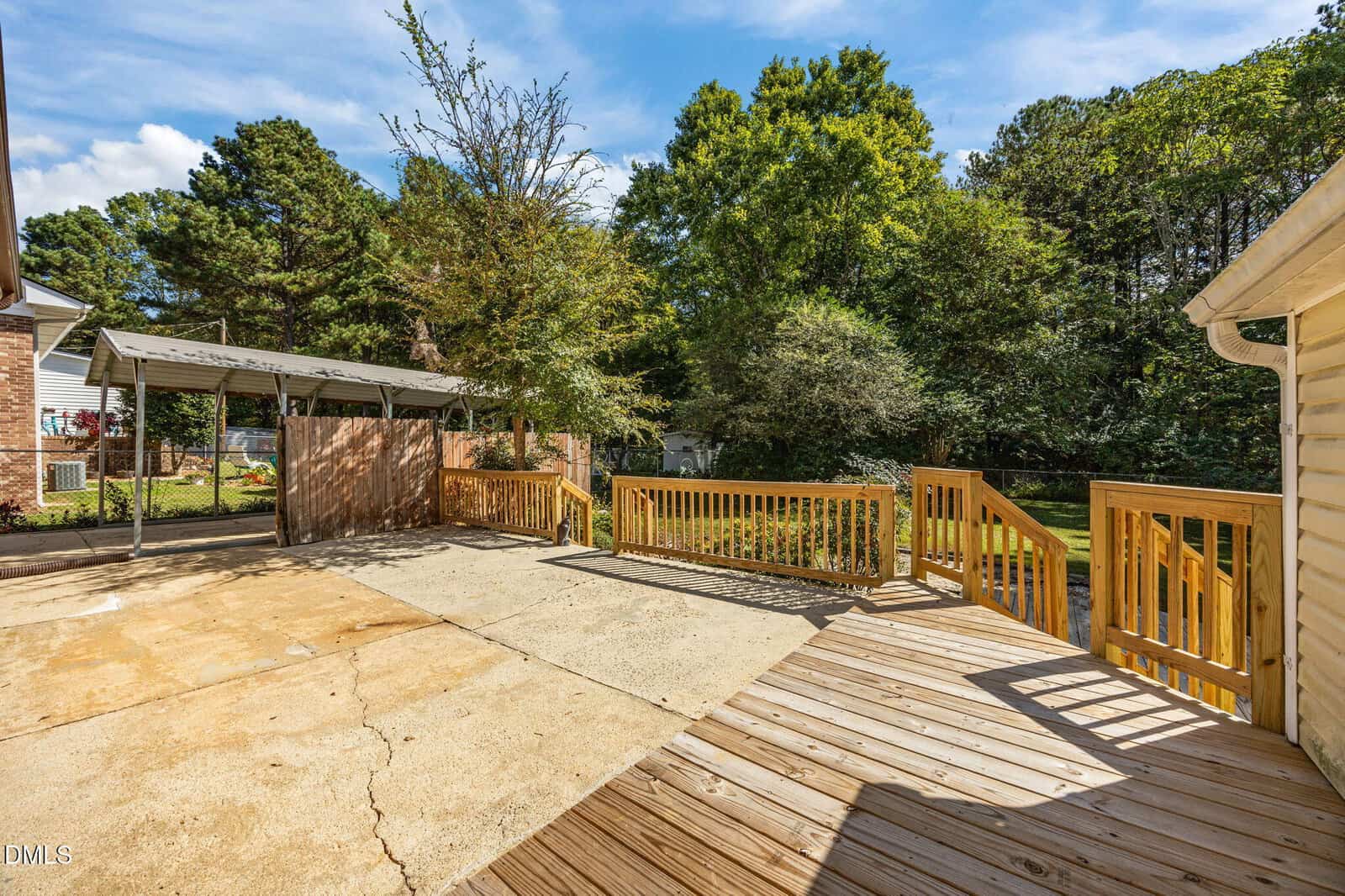 A backyard area with a concrete patio, a wooden deck with railings, a carport, and surrounding trees in the background on a sunny day.