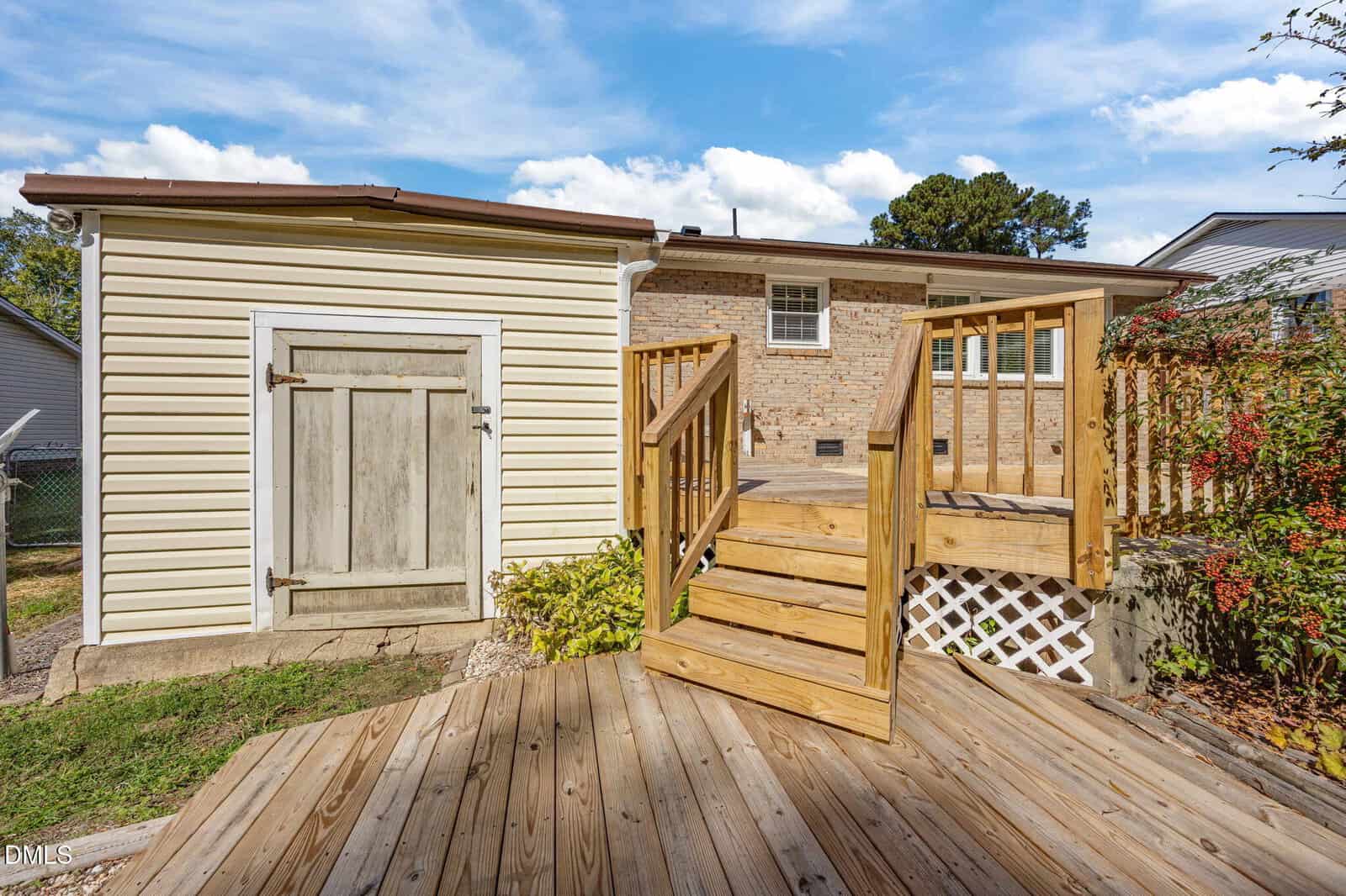 A wooden deck with stairs leads up to a raised entryway at the back of a brick house. There is a small shed or storage area with a weathered wooden door to the left of the deck. The sky is partly cloudy.