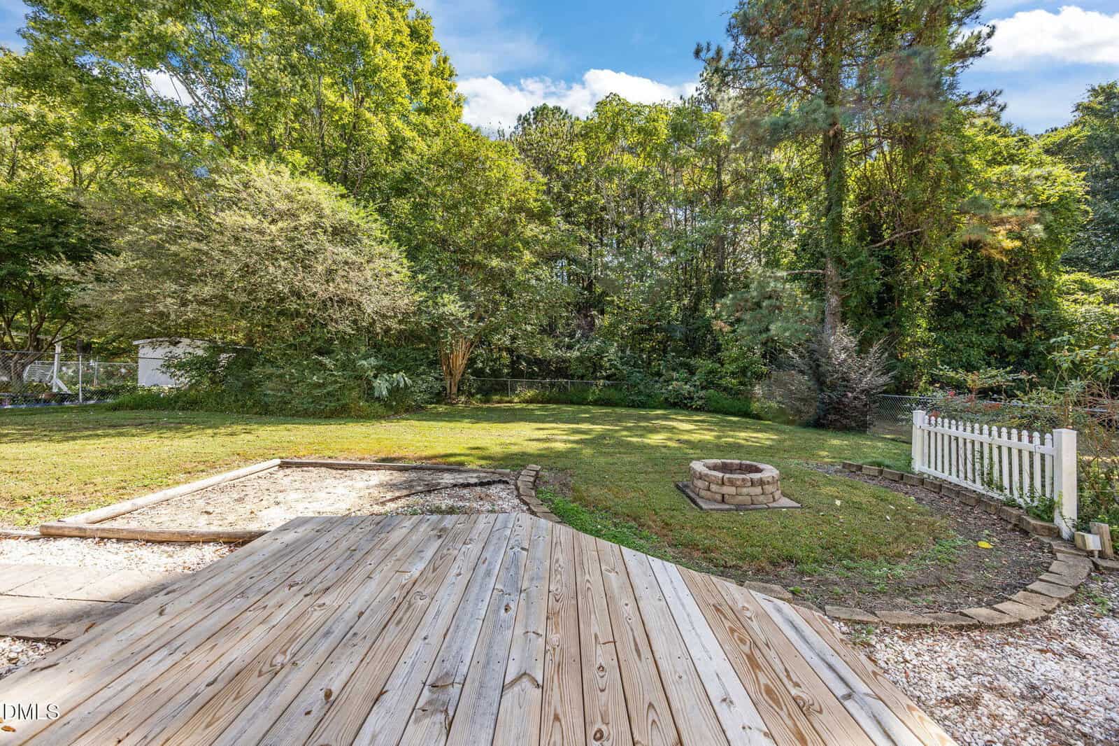 A backyard with a wooden deck in the foreground, a circular fire pit on a grassy lawn, a small white fence to the right, and dense trees and shrubs in the background under a blue sky with clouds.