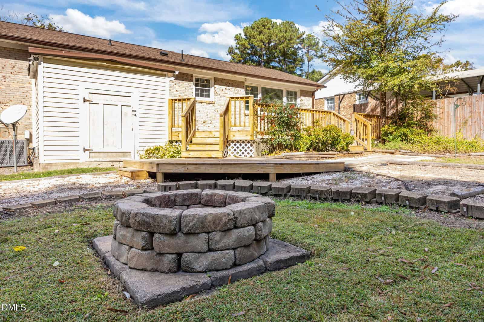 A backyard with a circular stone fire pit on grass, a wooden deck with steps, and a brick house in the background under a blue sky with scattered clouds.