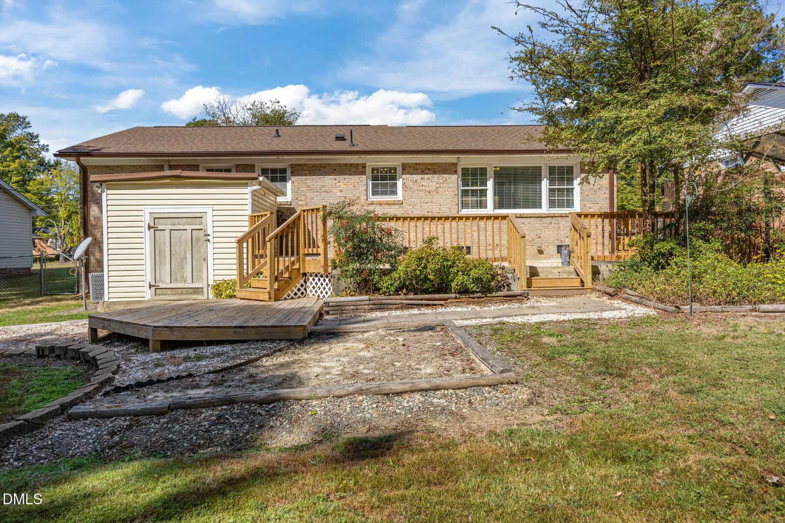 Single-story brick house with a wooden deck and ramp, surrounded by a grassy yard and trees. The sky is blue with scattered clouds, and there are garden beds near the deck.