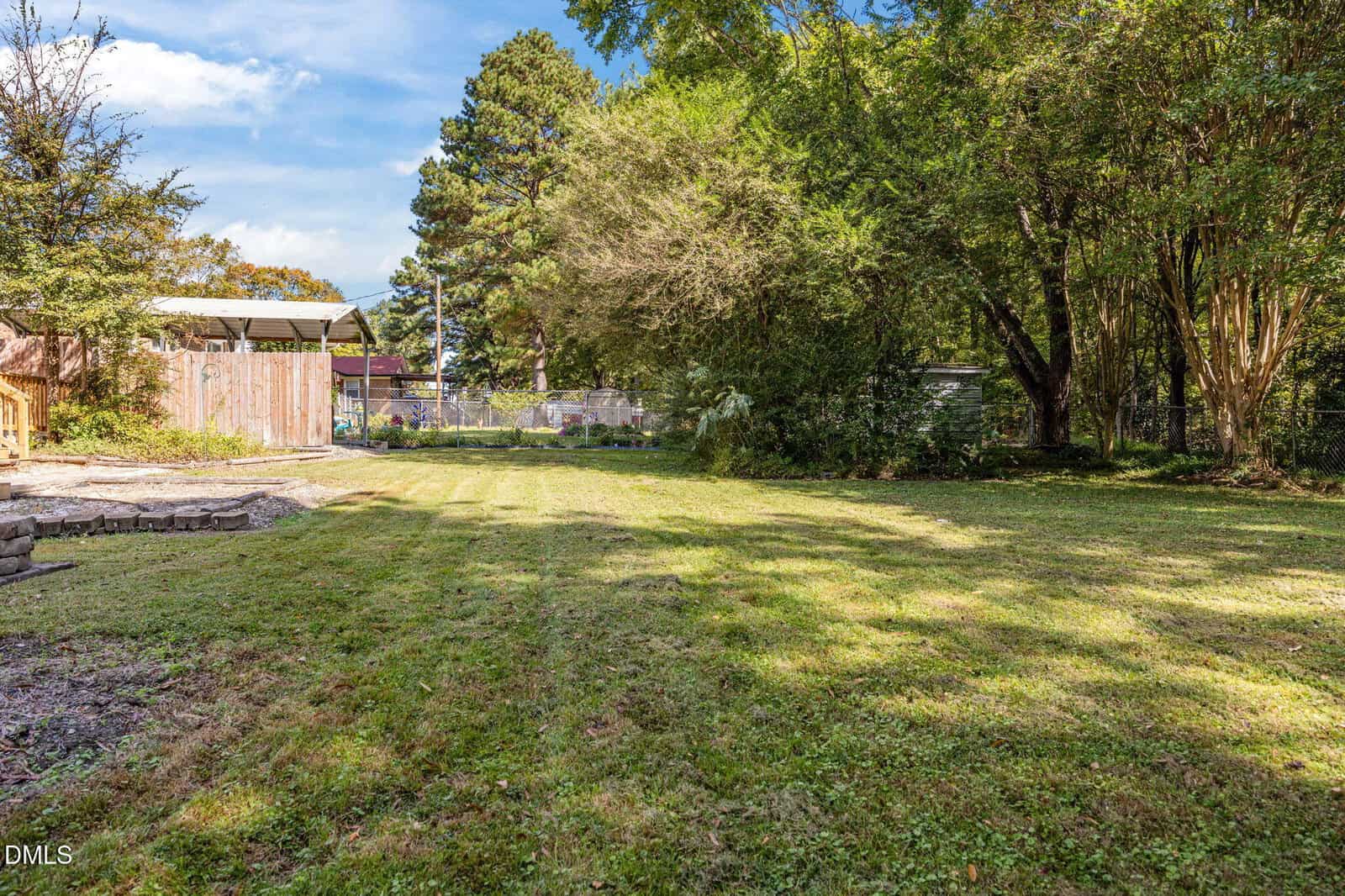A spacious backyard with green grass, bordered by trees and a wooden fence, under a blue sky with scattered clouds. There is a small rock border and some shaded areas from the trees.
