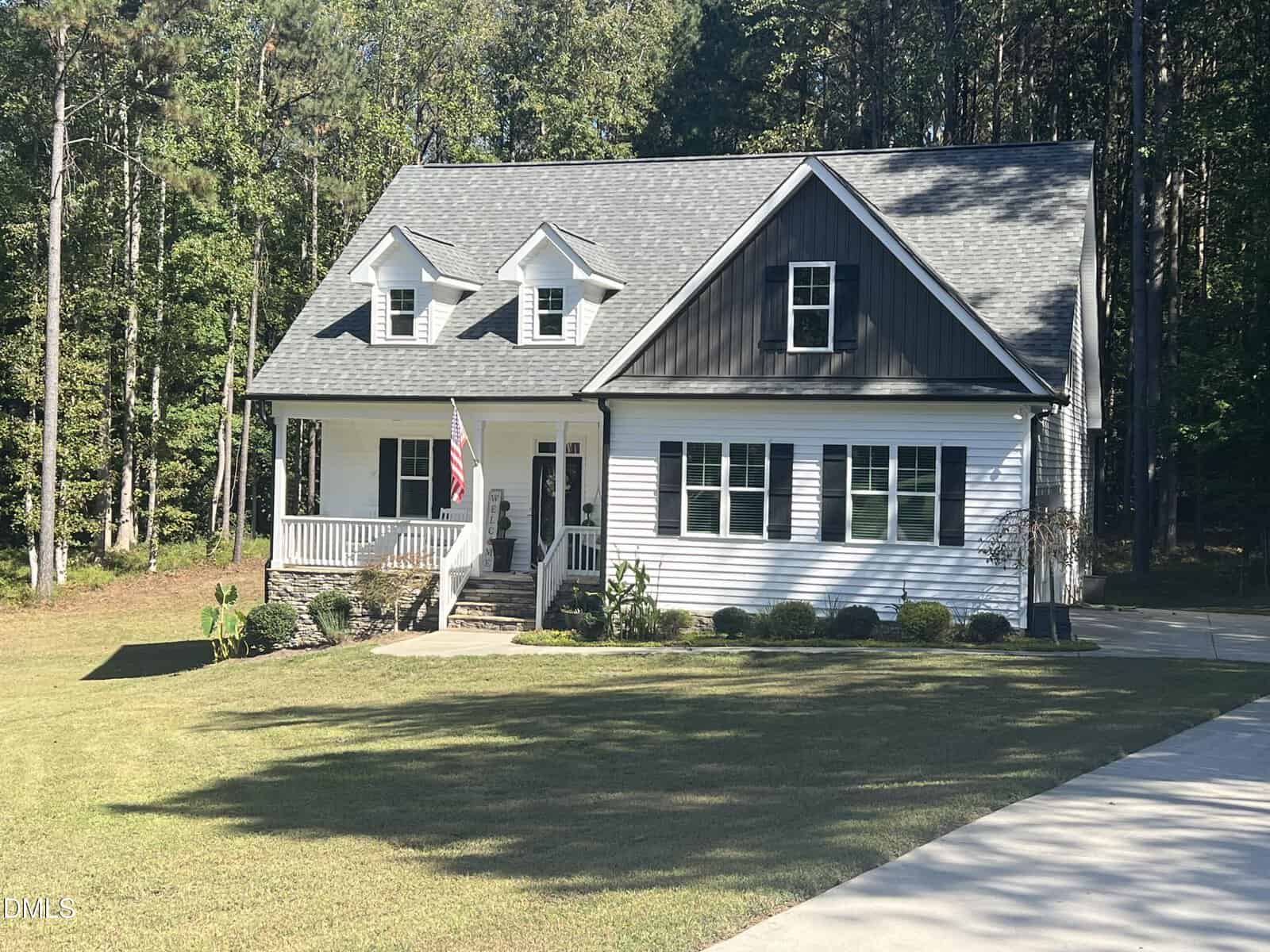 A white two-story house with a dark roof and dormer windows sits proudly at 220 Kingfisher Way, Louisburg. The front porch, adorned with an American flag, overlooks bushes along the front and a driveway leading up. Surrounded by a lush lawn and wooded trees in the background, this home offers classic charm in a serene setting.