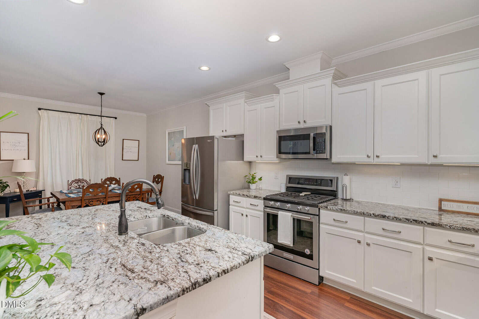 Modern kitchen with white cabinets, stainless steel appliances, granite countertops, and a central island with a sink; dining area with a wooden table and chairs is visible in the background.
