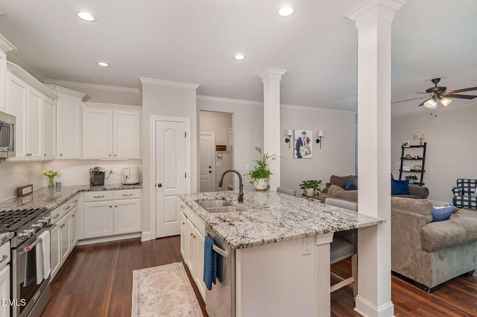 Open kitchen with white cabinets, stainless appliances, granite countertops, and an island with a sink. Adjacent living area with a gray sofa, armchair, shelves, and ceiling fan. Hardwood floors throughout.