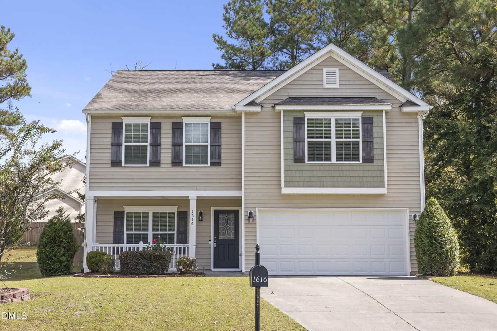 A two-story suburban house in Creedmoor with beige siding, black shutters, a small front porch, double garage, and concrete driveway. Shrubs and trees surround the home at 1616 Irving Place, with a house number sign by the drive.