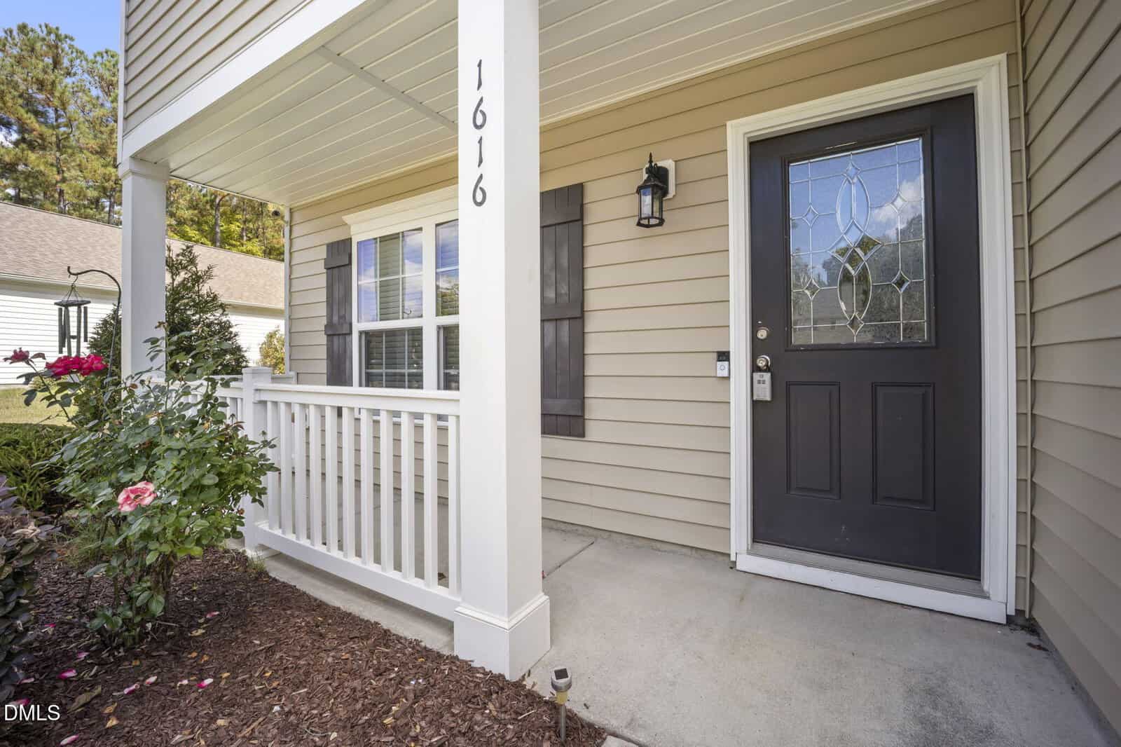 A small covered front porch with a white railing, tan siding, a black front door with decorative glass, house number 1616, a light fixture, window with shutters, and flower bed with blooming roses.