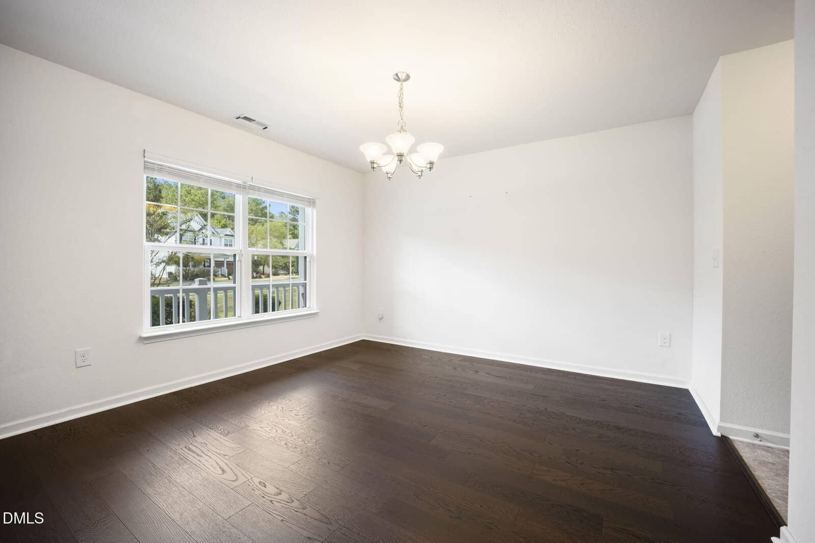 Bright, empty room with dark hardwood floors, white walls, a large double window, and a ceiling light fixture. Natural light comes through the window, and greenery is visible outside.