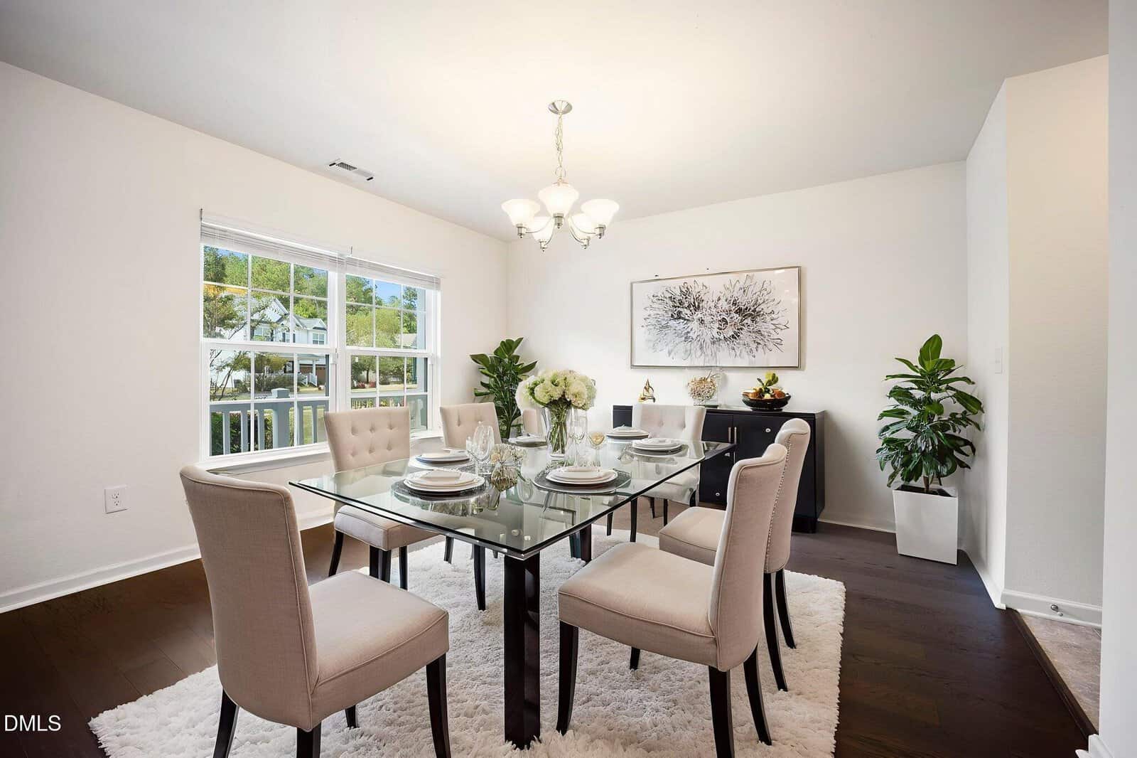 A dining room with a glass table set for six, beige upholstered chairs, a white rug, dark wood floors, white walls, potted plants, a sideboard, and a chandelier. Large window lets in natural light.