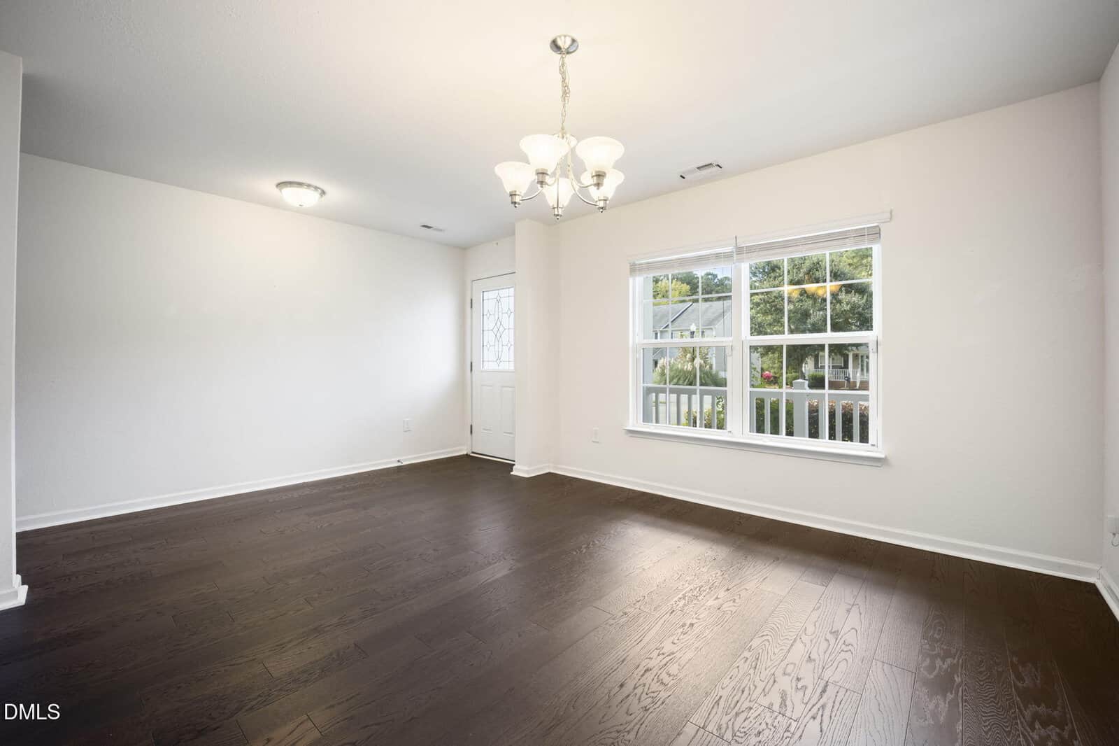 Bright empty room with dark hardwood floors, white walls, large window showing greenery outside, chandelier ceiling light, and a white door with a small window near the corner.
