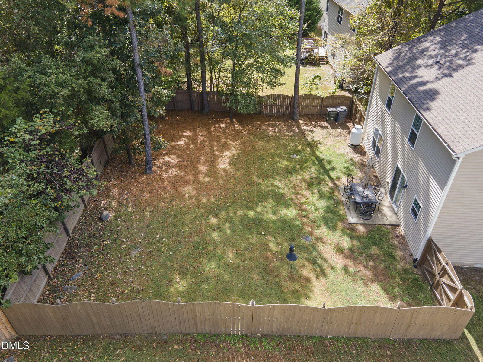 A fenced backyard with a mix of grass and scattered fallen leaves, bordered by trees. A patio with outdoor furniture and a grill is adjacent to a beige two-story house on the right side.