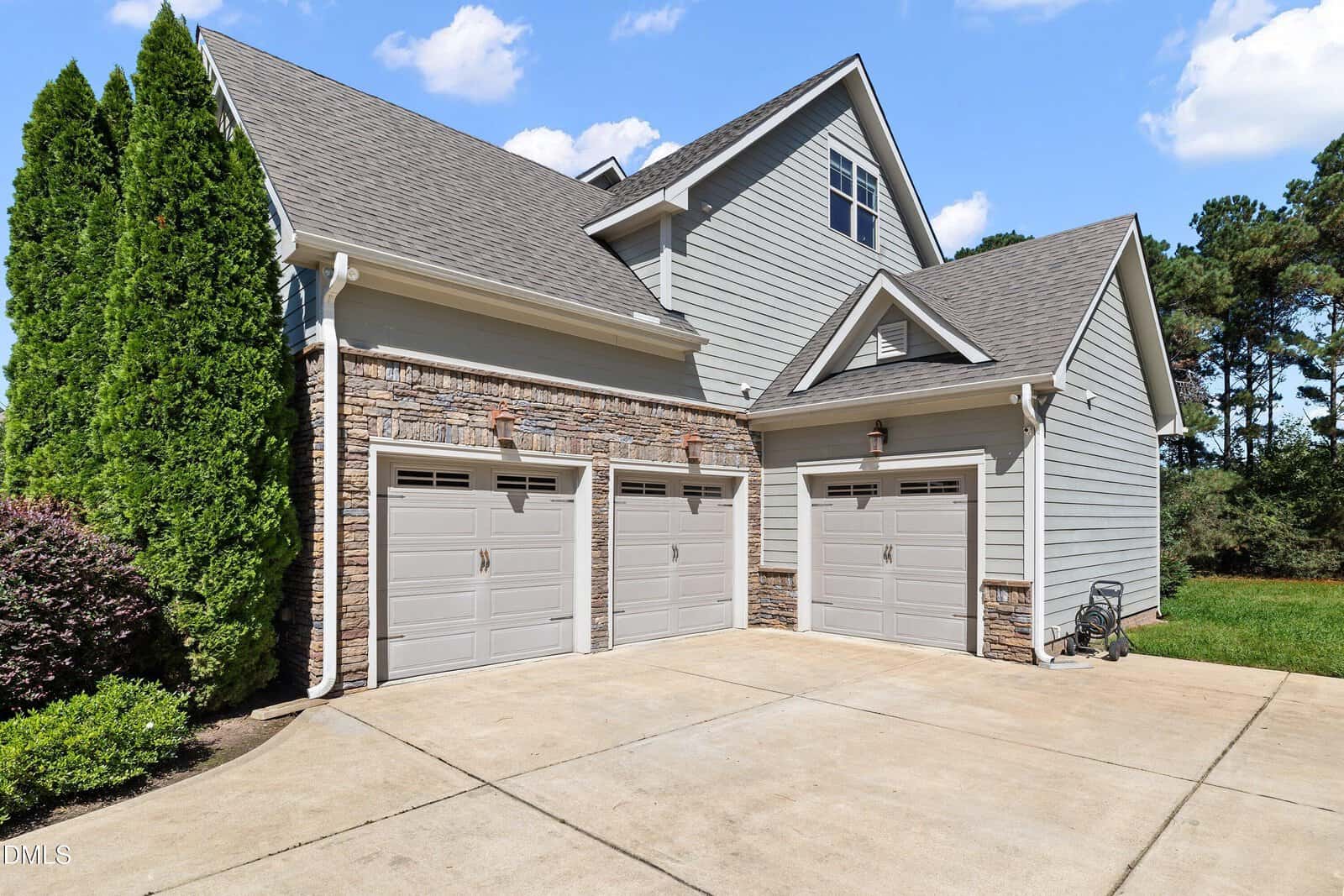 A three-car garage attached to a modern house with gray siding, stone accents, and a concrete driveway. Bushes and trees are visible on the left and right sides, with a green lawn in the background.
