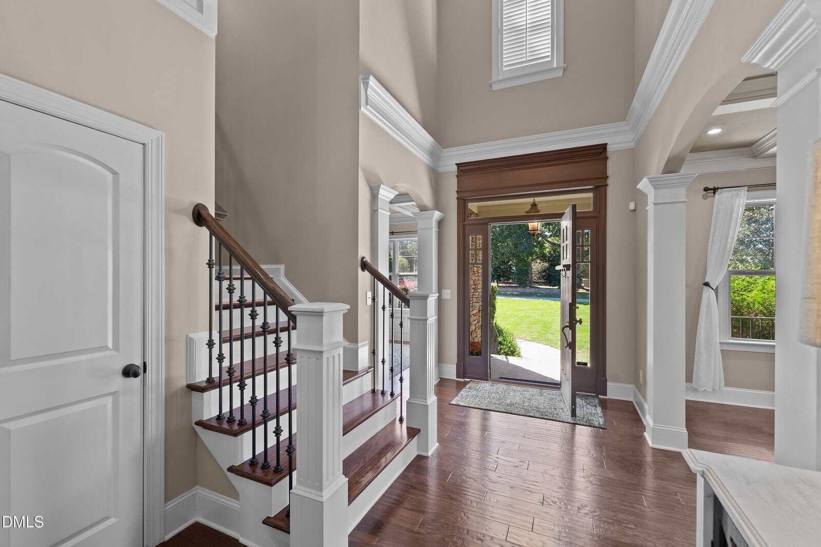 A spacious entryway with hardwood floors, a wooden staircase with white trim and black balusters, tall ceilings, and large windows allowing natural light. An open front door leads to a green lawn outside.