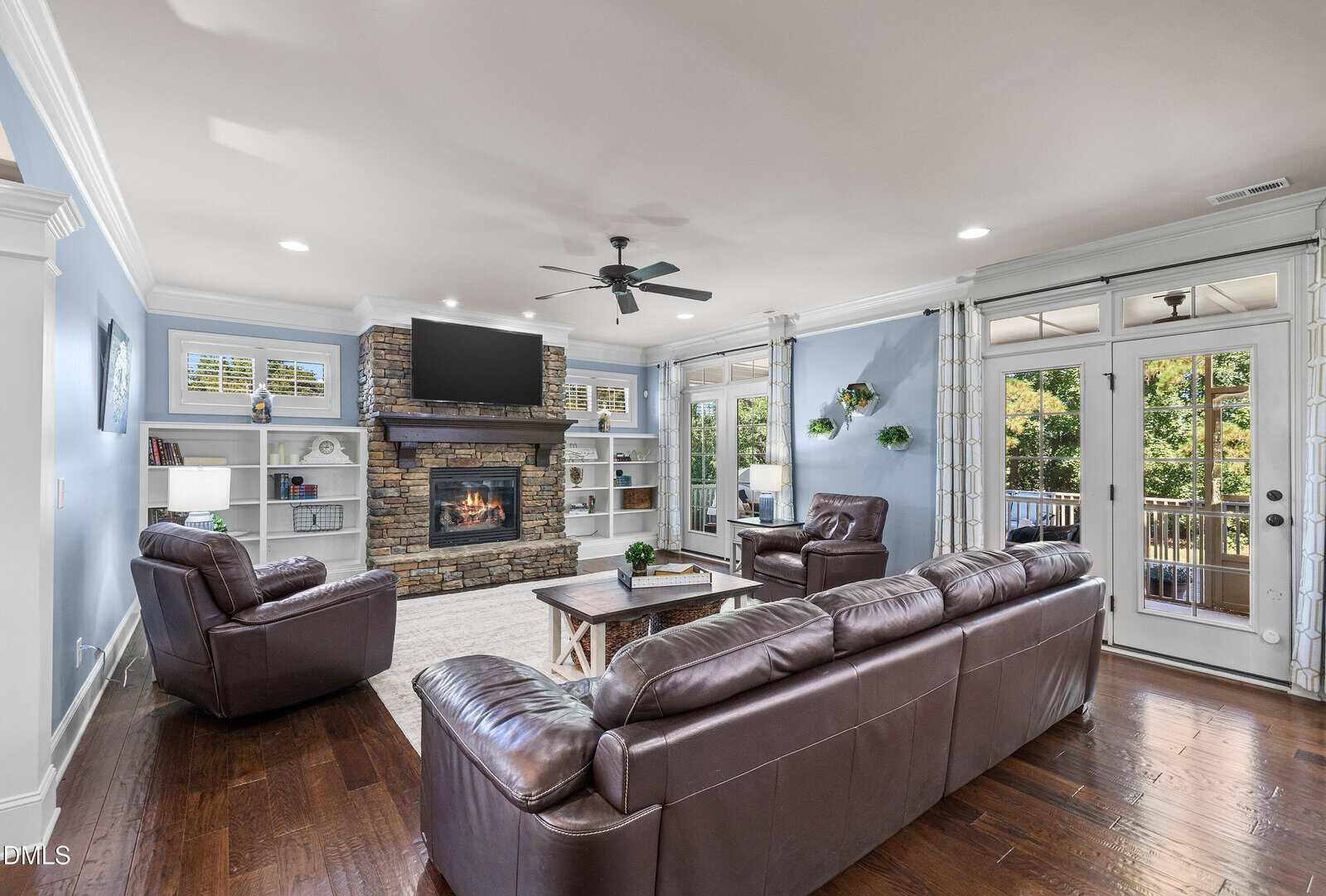 Spacious living room with brown leather sofas, stone fireplace, wall-mounted TV, built-in shelves, hardwood floors, blue walls, ceiling fan, and large windows with a view of a deck and trees outside.