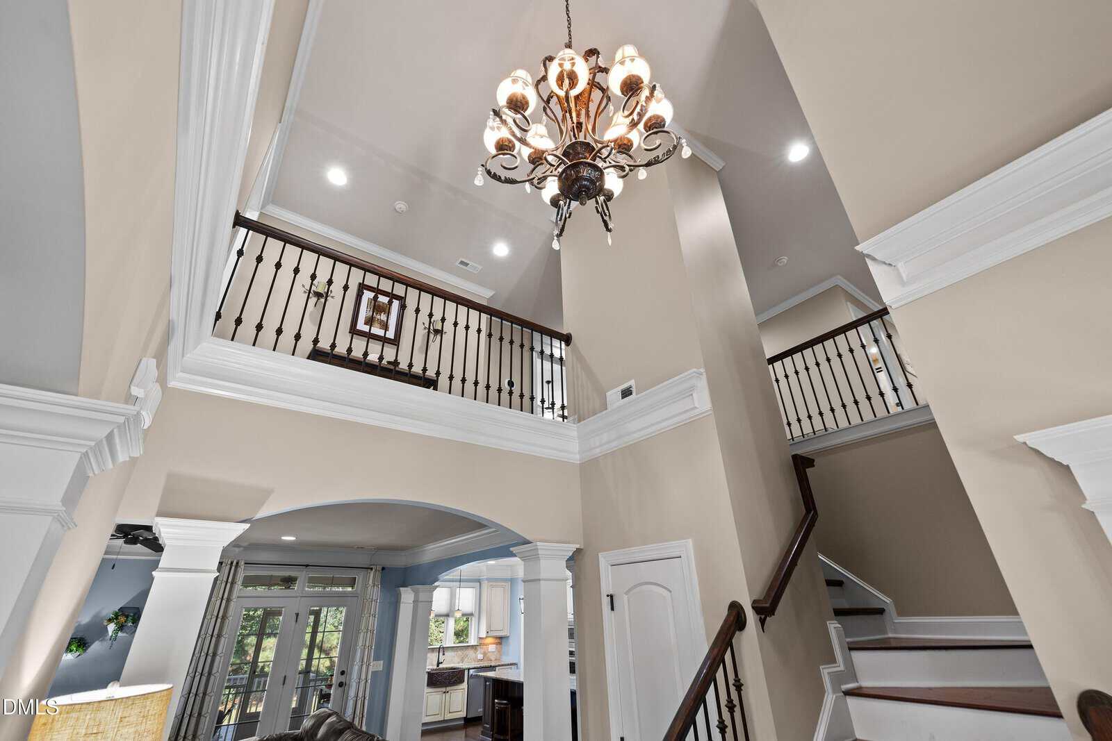 A view of the two-story home at 690 Hawthorne Place features a staircase, balcony with black railings, beige walls, white trim, and a decorative chandelier. The kitchen and large windows are visible in the background.
