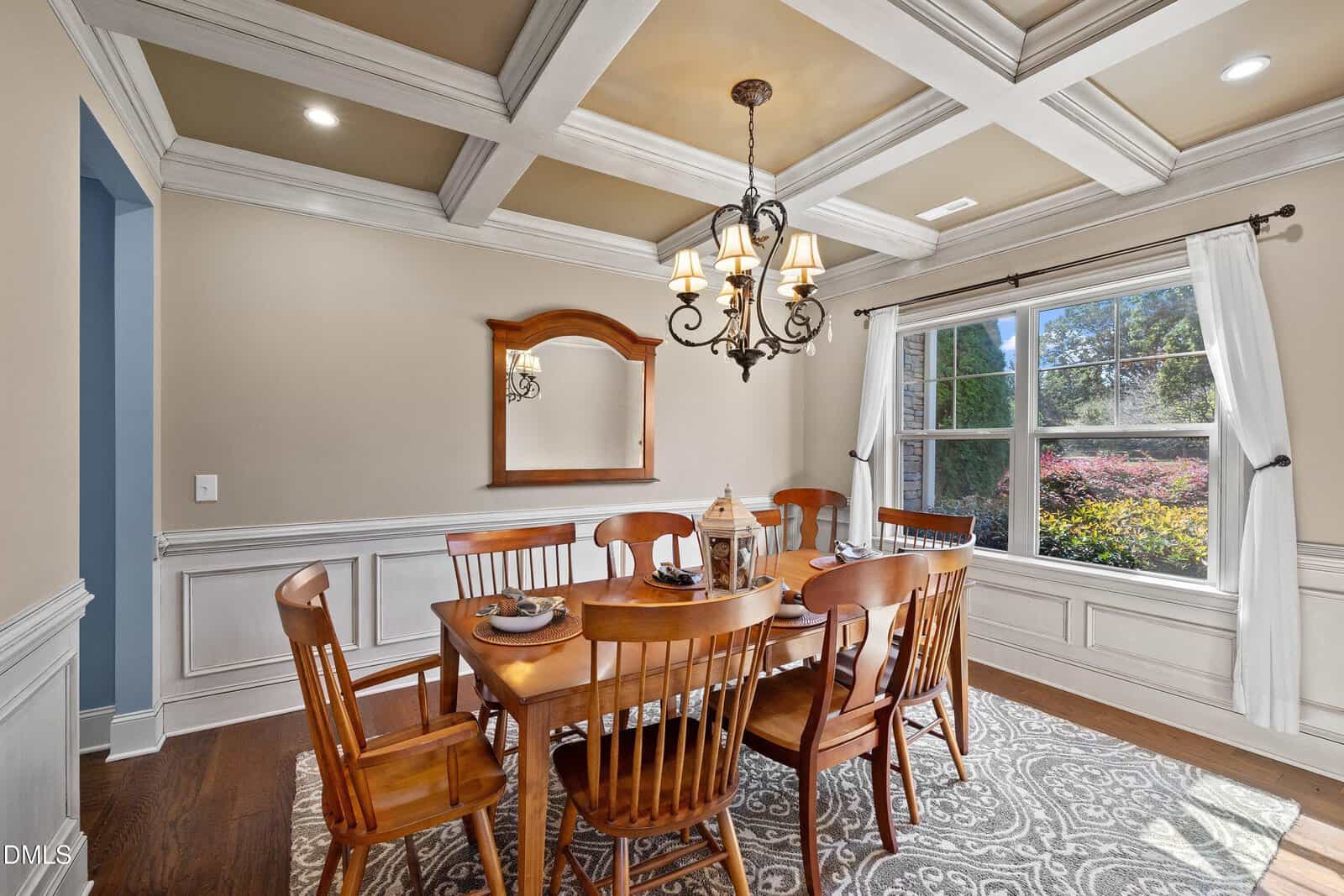 Elegant dining room with a wooden table and six chairs, chandelier above, large window with white curtains, wall mirror, beige walls, and decorative ceiling beams. Natural light fills the room.
