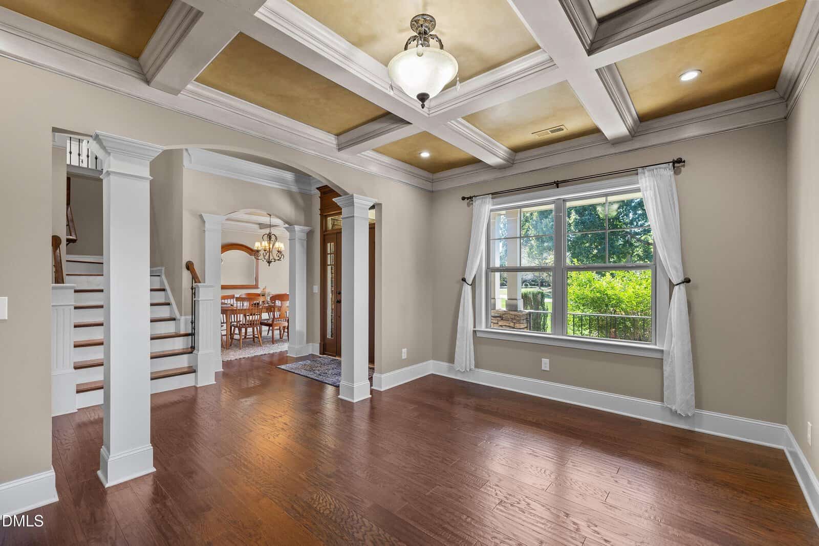 Spacious living room with coffered ceiling, hardwood floors, large window with white curtains, and view of adjacent dining room. Staircase visible to the left. Warm neutral color palette.