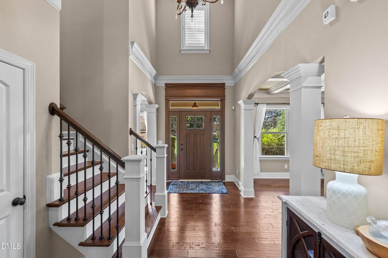 A spacious foyer with a wooden front door featuring glass panels, a staircase with white railings, hardwood floors, a patterned rug, and neutral-colored walls. Natural light enters through windows above and beside the door.