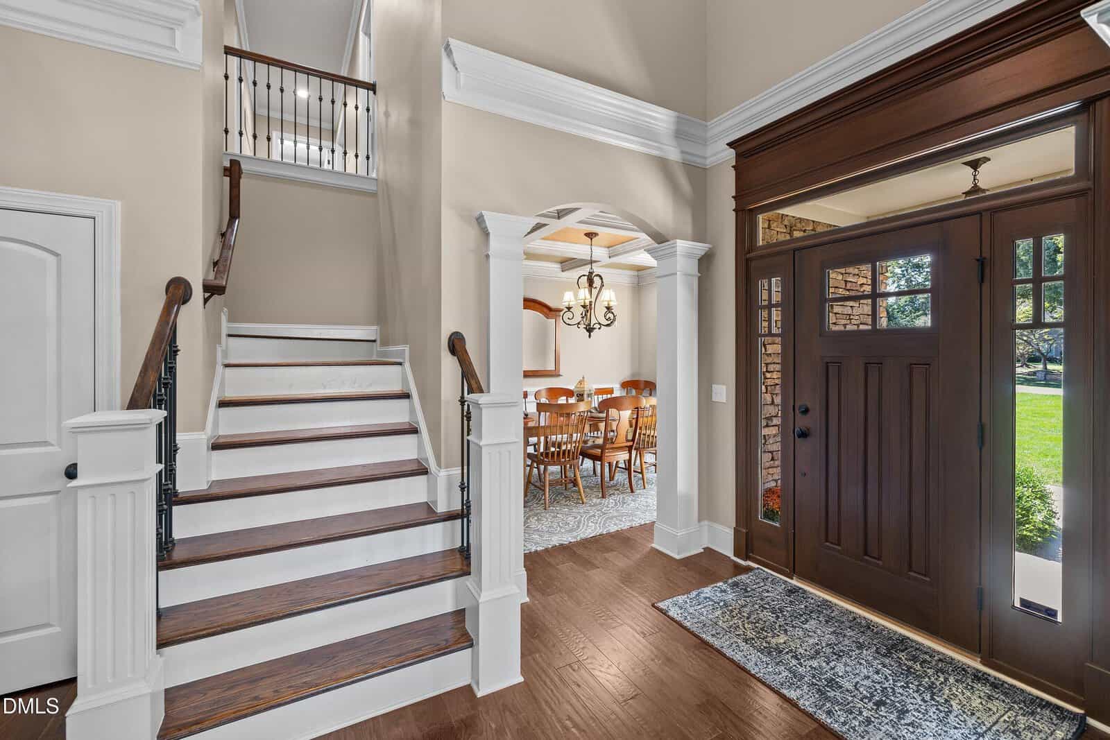 A view of a home's foyer with a dark wooden front door, staircase with wooden steps and white risers, open doorway to a dining room with a chandelier and wooden table, and large windows letting in natural light.