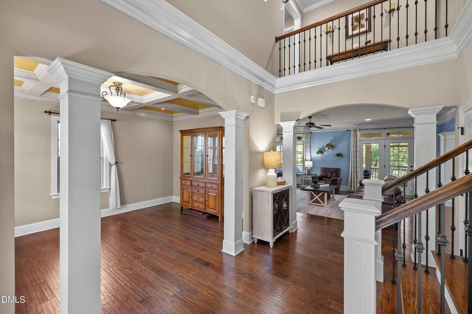 Open foyer with hardwood floors, white columns, and crown molding. A wooden staircase with a black railing leads upstairs. Dining and living rooms are visible, both furnished with wood pieces and neutral decor. Natural light fills the space.