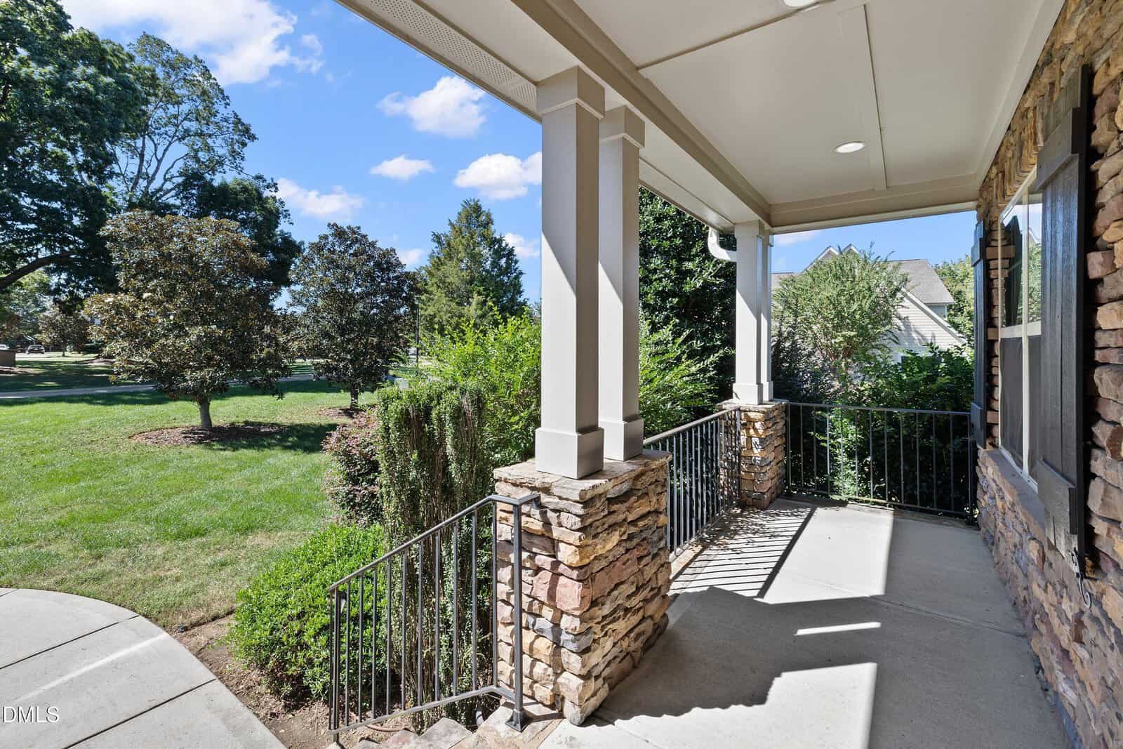 A covered front porch with stone pillars and metal railings overlooks a landscaped yard with green grass, shrubs, and trees under a blue sky with scattered clouds.