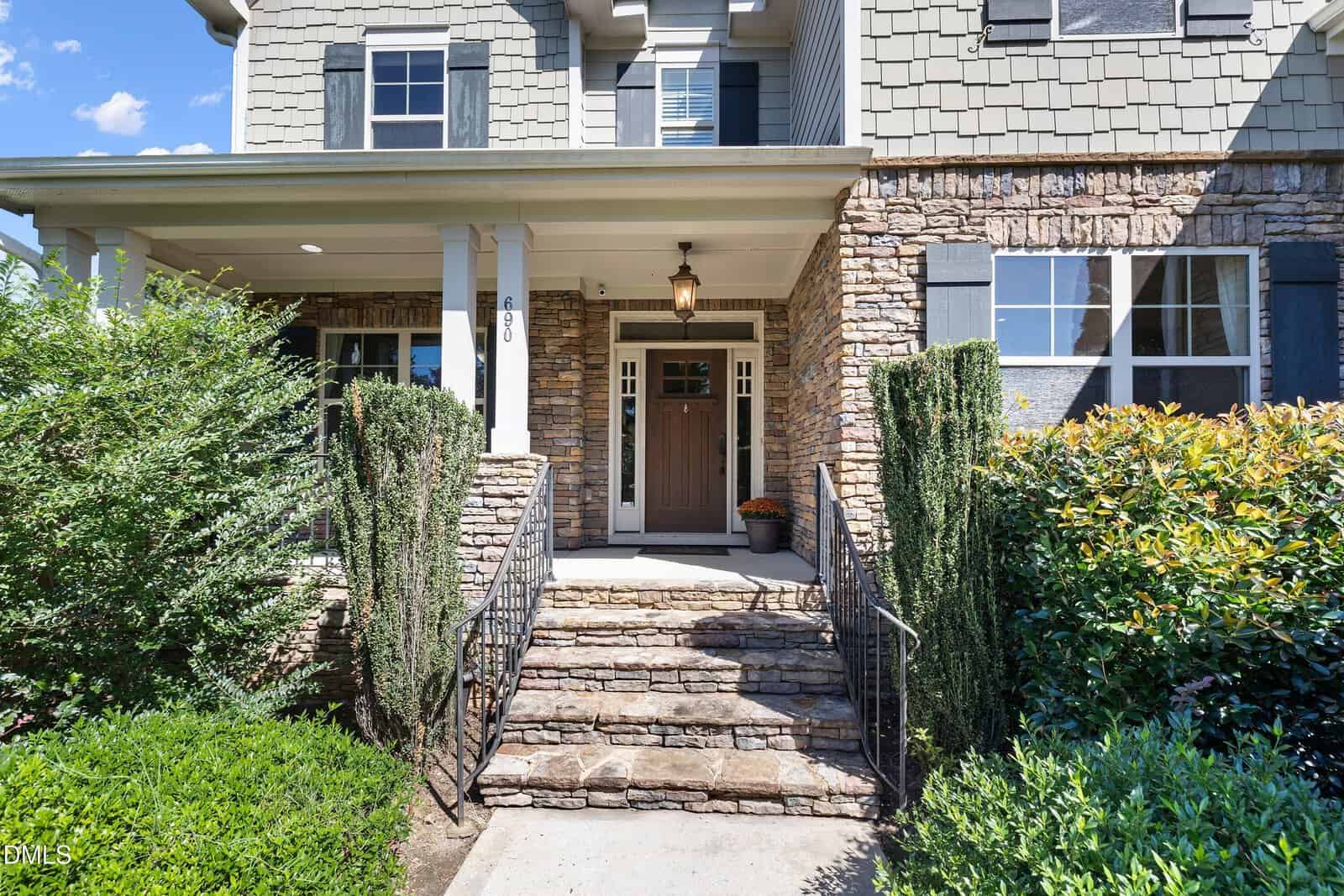 A stone walkway with steps leads to a covered front porch of a house with a dark brown door, sidelights, two white pillars, and stone and shingle exterior walls. Shrubs and bushes line the entrance.