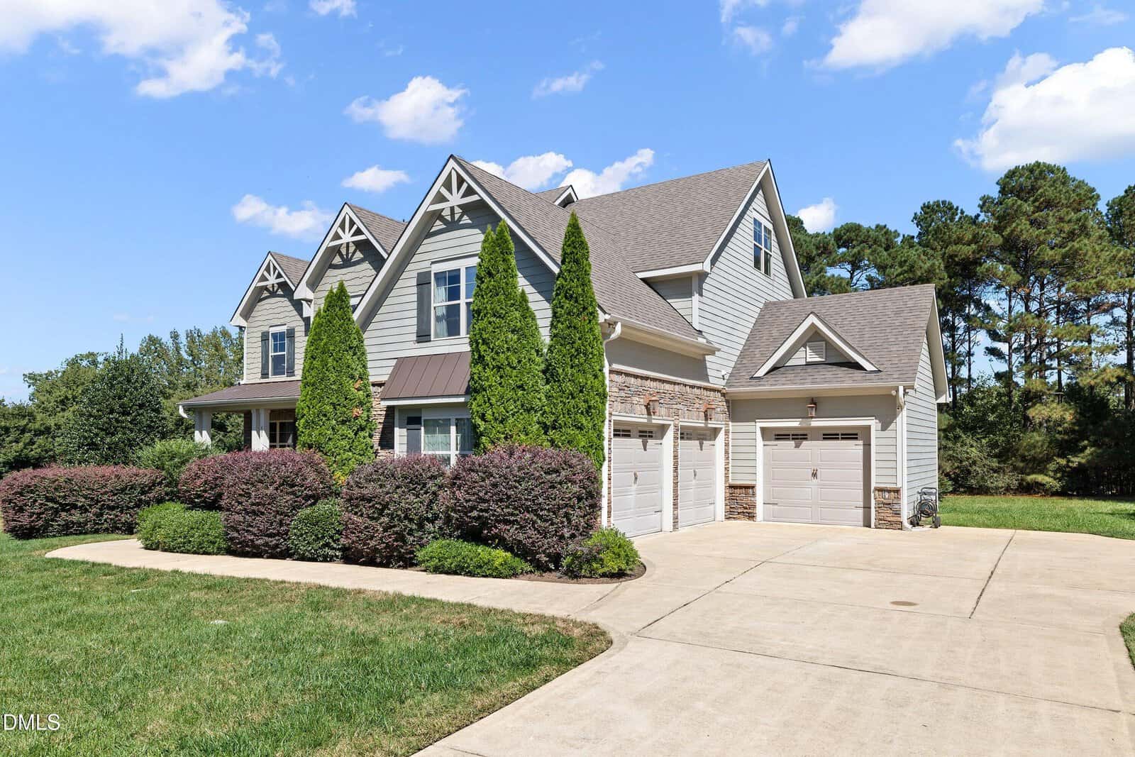 Two-story suburban house with gray siding, a gabled roof, three garage doors, and landscaped bushes and trees in front, set against a blue sky with scattered clouds.