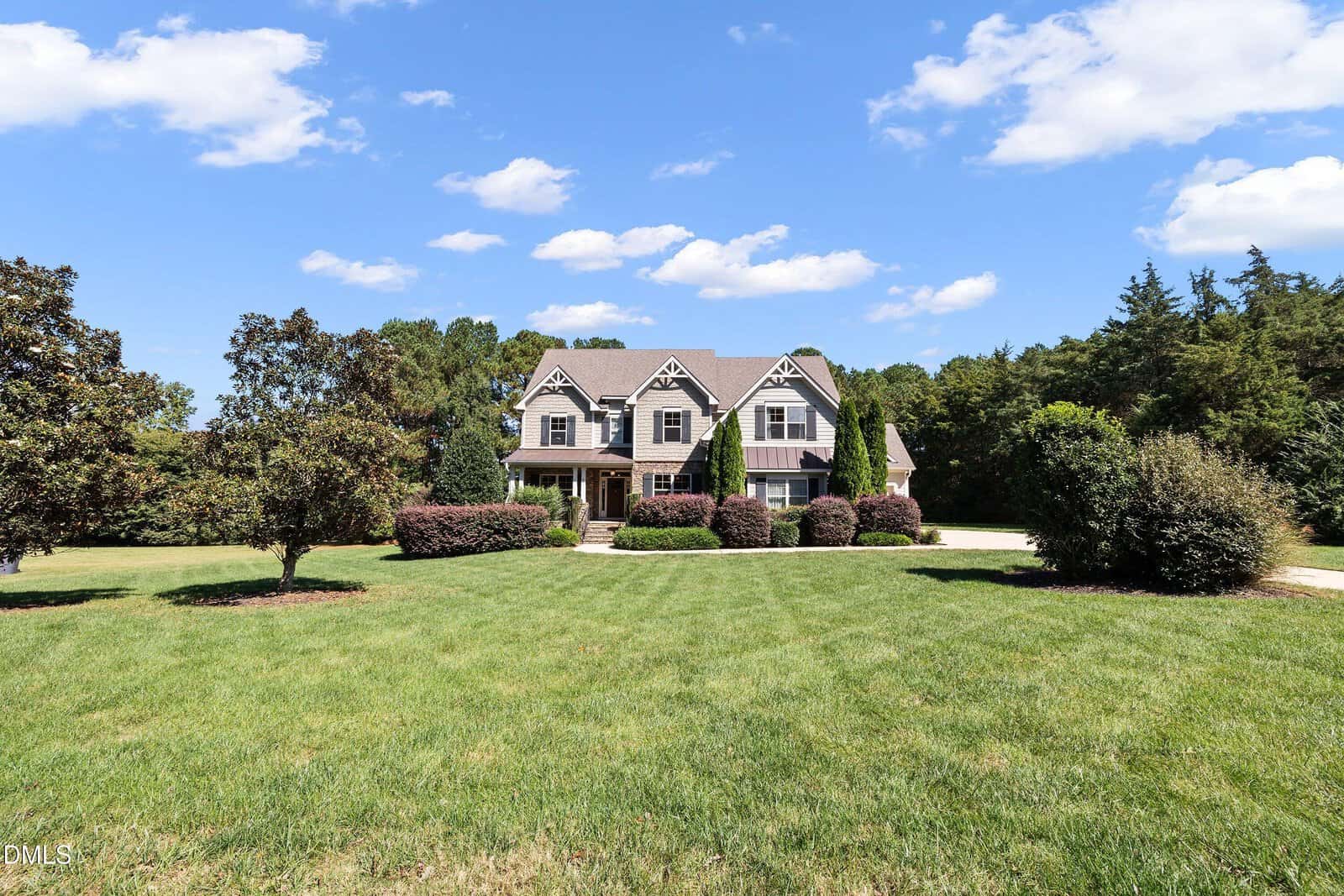 A two-story house with a covered porch sits on a large, well-maintained lawn. The house features gables, dormer windows, and is surrounded by shrubs and trees under a blue sky with clouds.