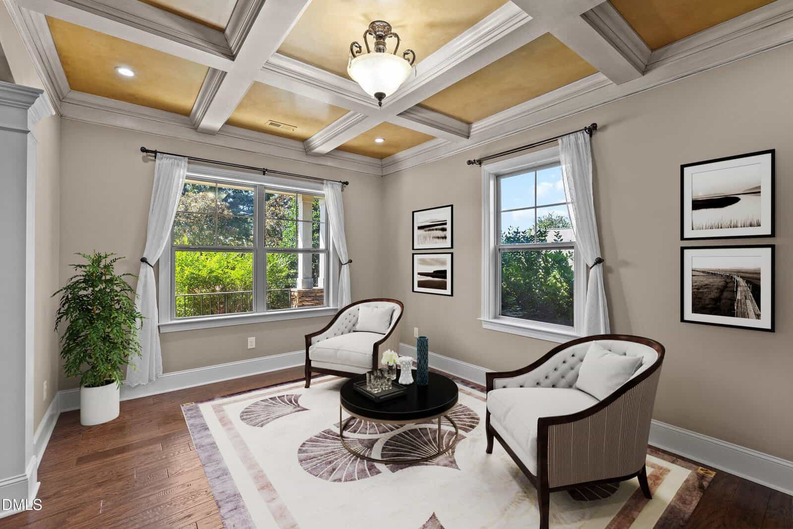 A small sitting room with two white armchairs, a round black coffee table, framed art on the wall, a large window, wood flooring, a potted plant, and a tray ceiling with a central light fixture.