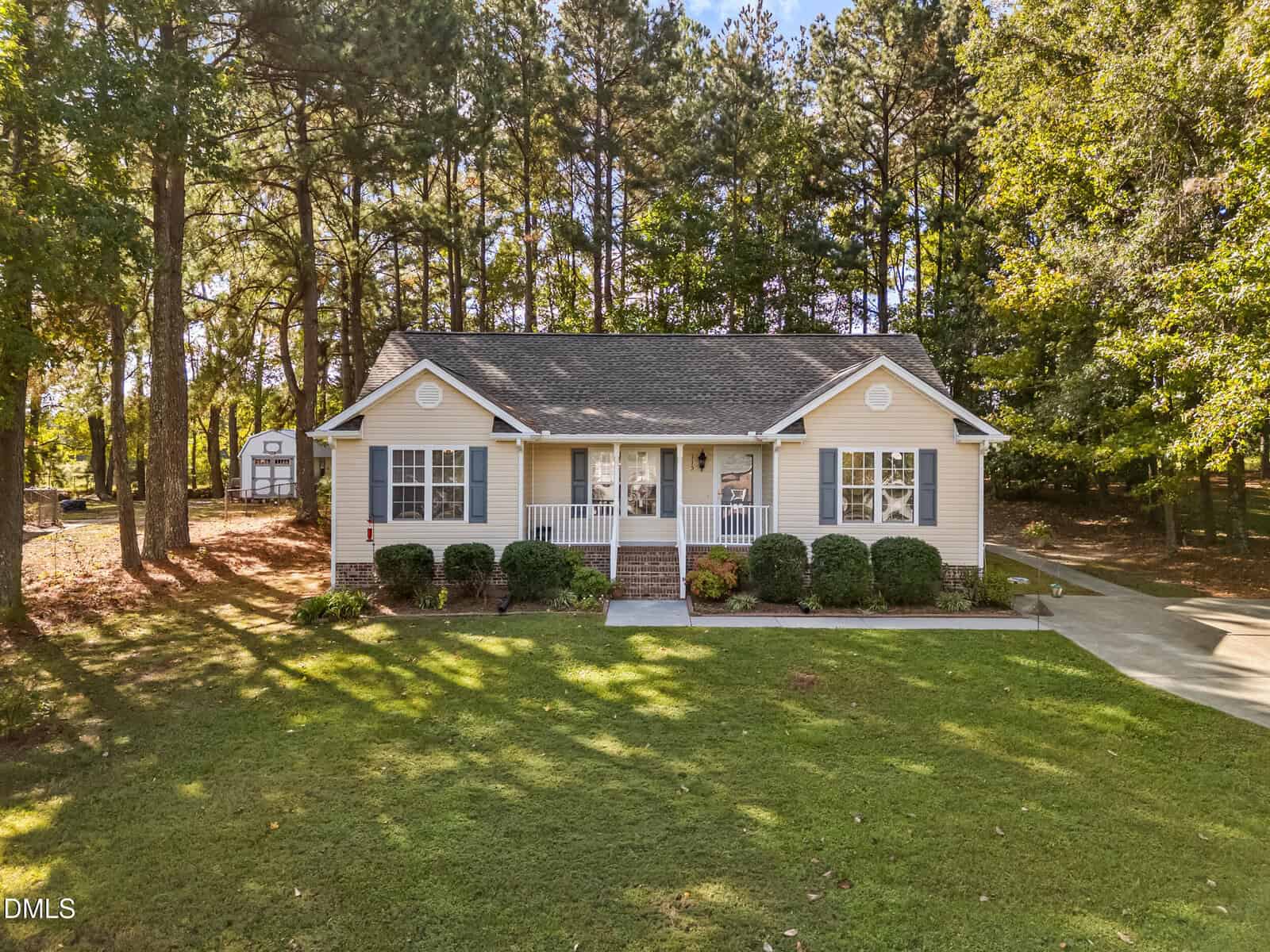 A single-story beige house with white trim, a covered front porch, and steps leading to the entrance. The house is surrounded by green lawn, bushes, and trees, with a driveway on the right and a shed in the background.