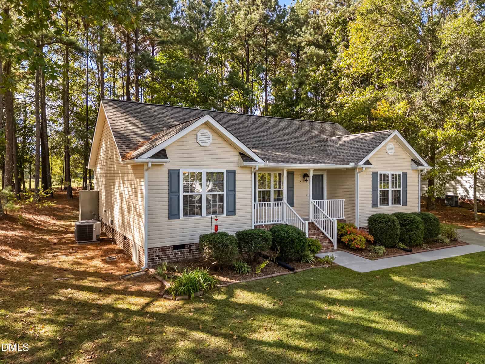 Single-story beige house with blue shutters, white trim, and a covered front porch. Surrounded by trees and shrubs, the home features a concrete driveway and a neatly maintained front lawn.