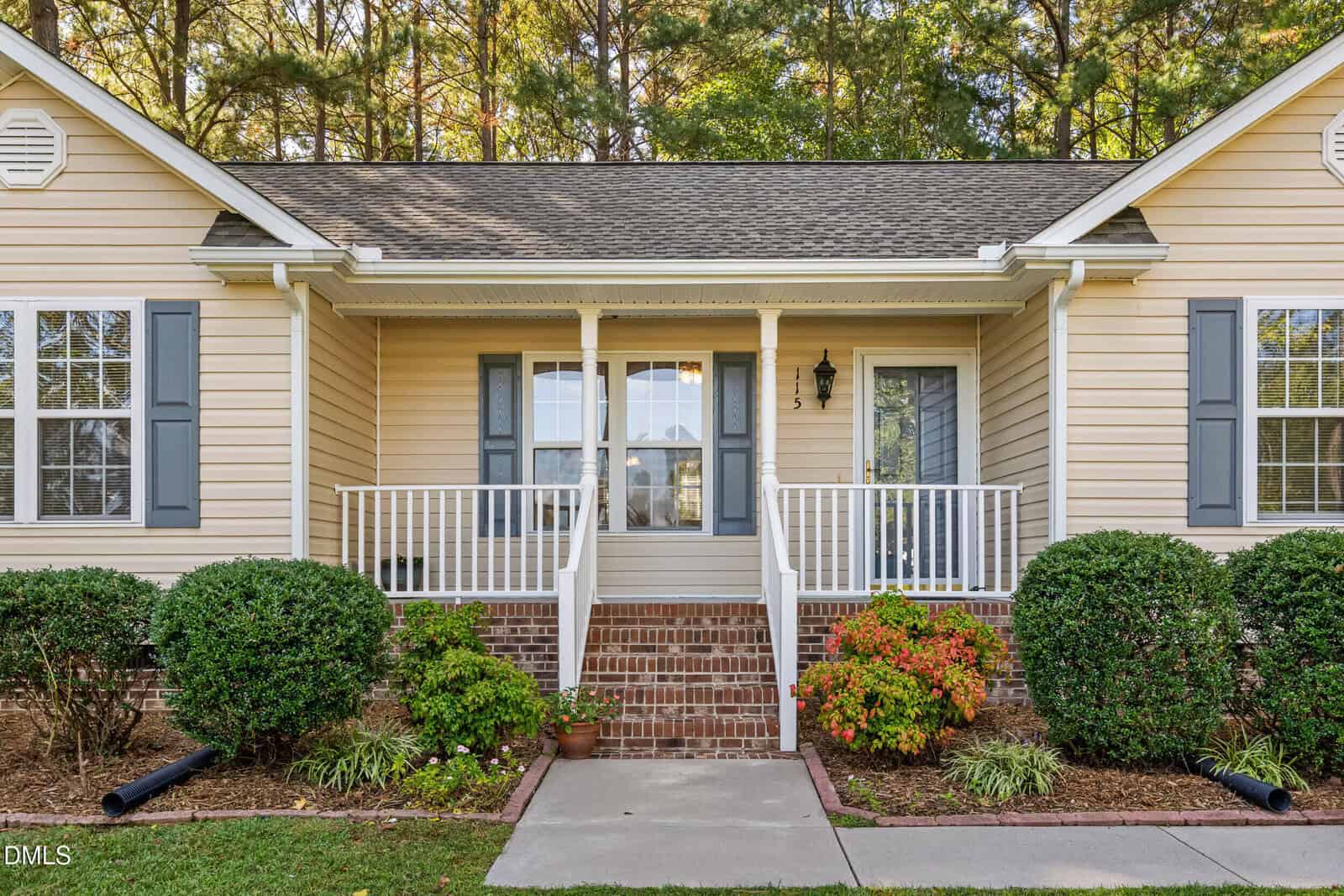 A beige single-story house with blue shutters, a covered front porch, white railings, brick steps, and neatly trimmed bushes and plants in the landscaped front yard.