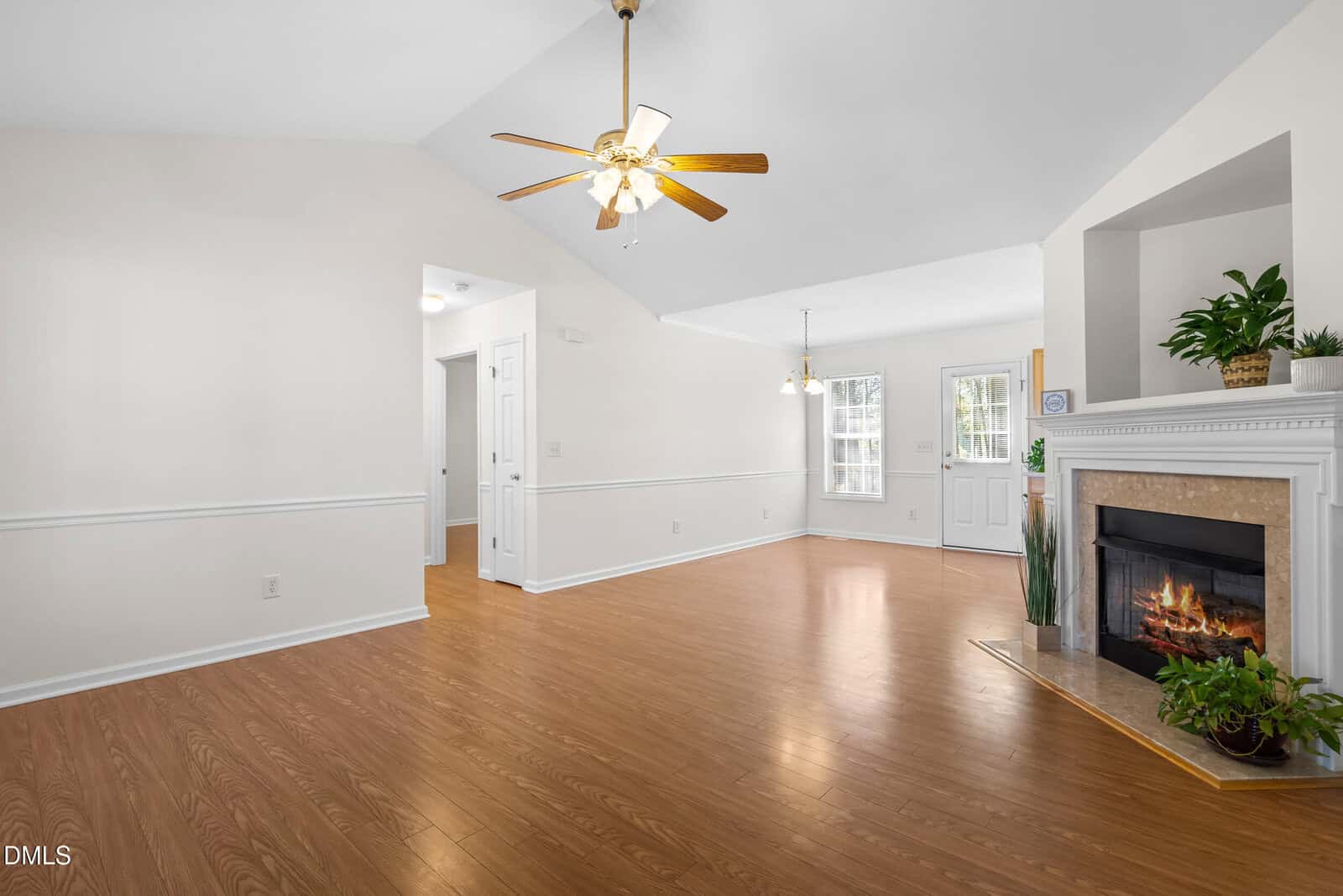 A spacious living room with wood flooring, vaulted ceiling, ceiling fan with lights, a white fireplace with a burning fire, potted plants on the mantel, and a view of the entryway and dining area.