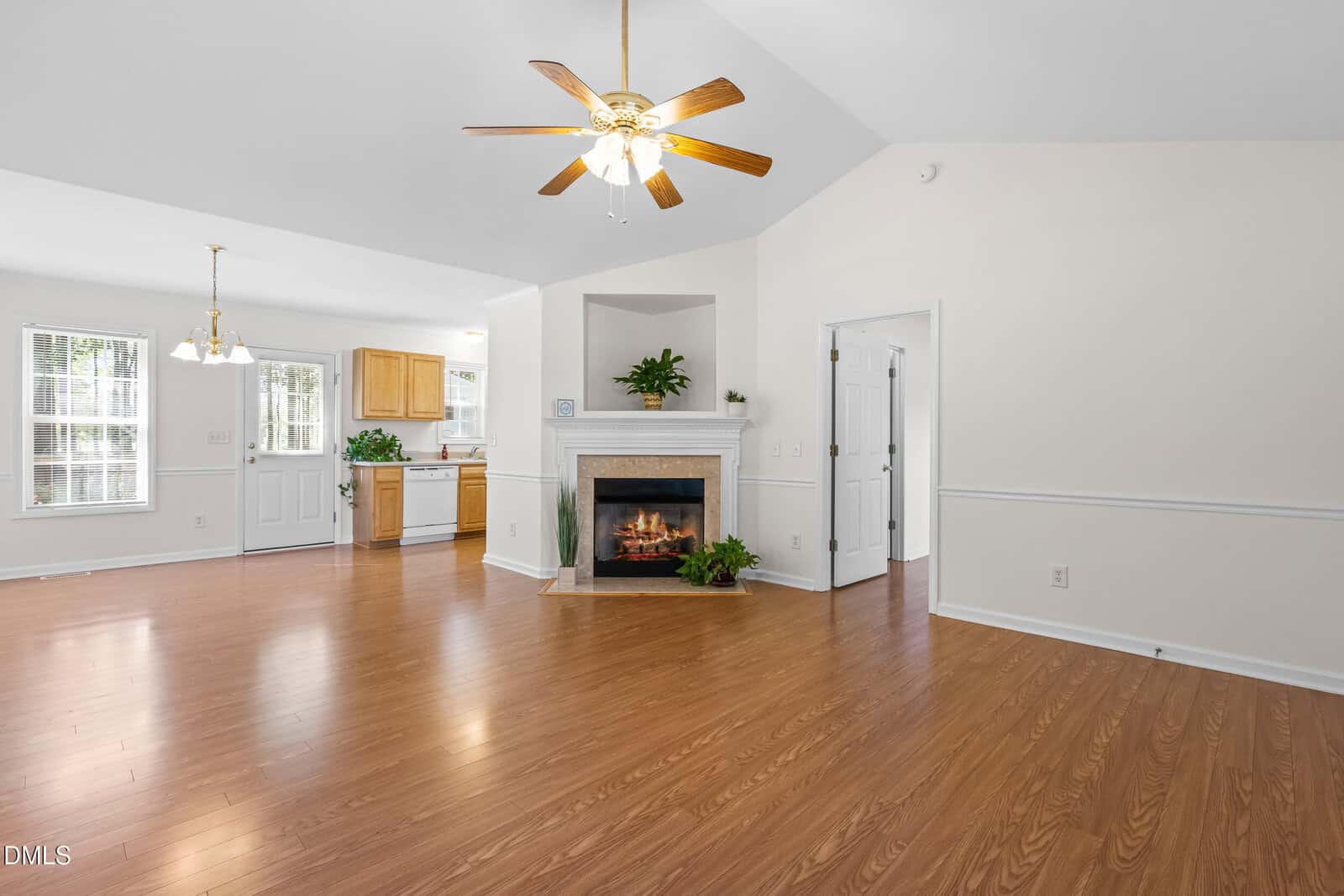 A spacious living area with wood flooring, a ceiling fan with lights, a white fireplace, and an open view into a kitchen with light wood cabinets. Large windows provide natural light.