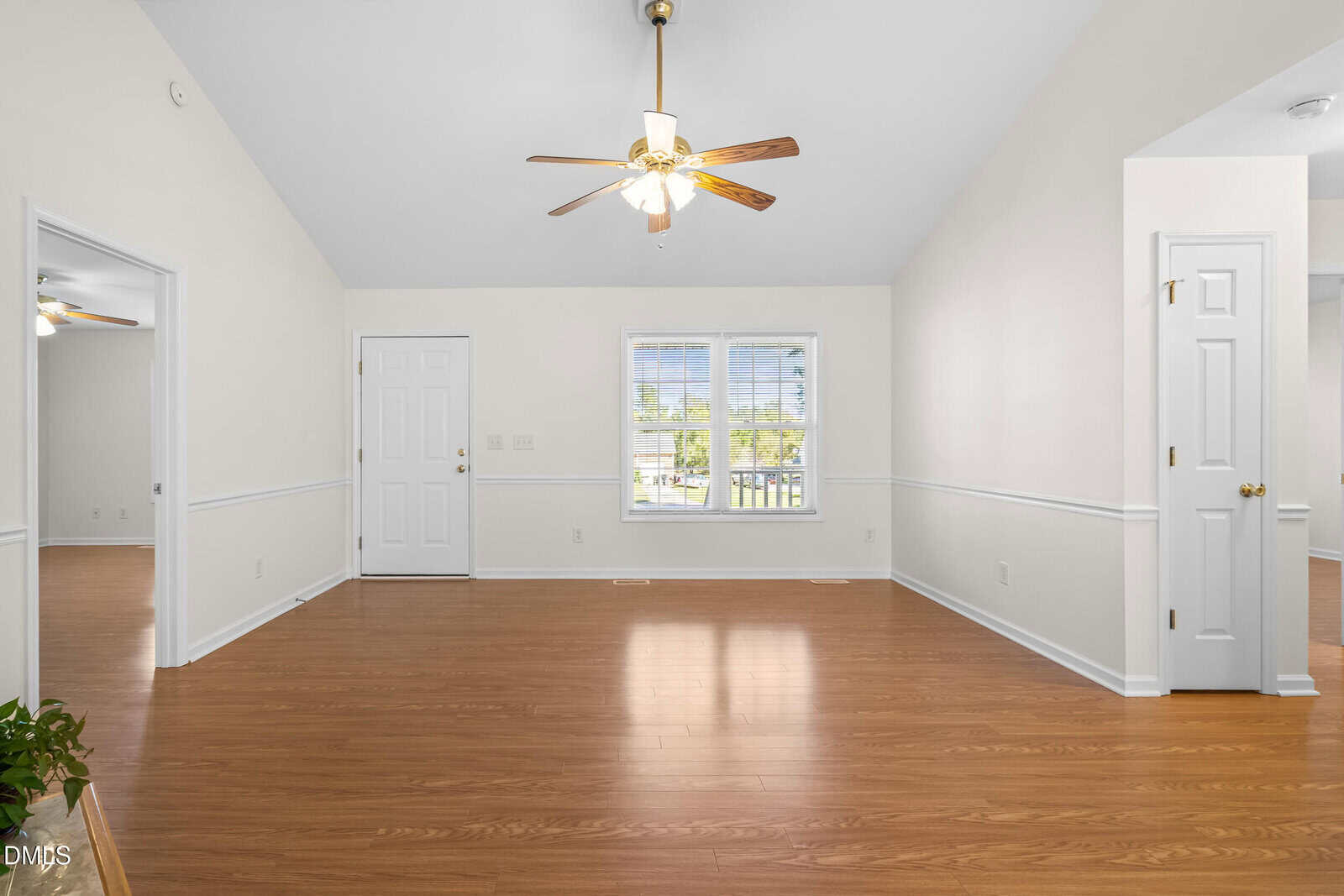 Bright, empty living room with wood floors, white walls, ceiling fan, window, and two white doors. A plant is partly visible in the left foreground.