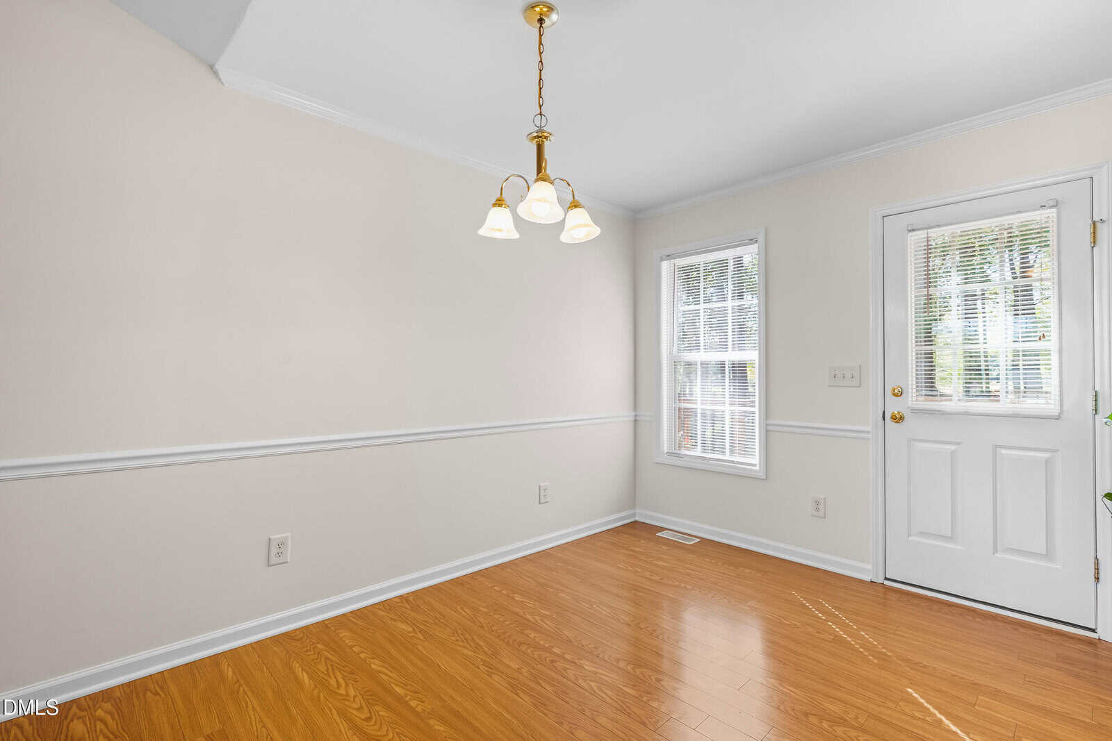 A small, empty room with light beige walls, wood flooring, a white door with glass panes, two windows with blinds, and a ceiling light fixture with three bulbs.