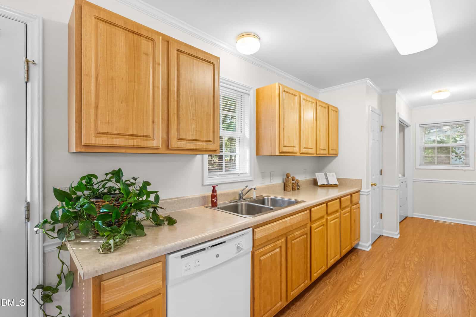 A kitchen with light wood cabinets, beige countertops, a double sink, a dishwasher, a houseplant, and a window above the sink. The room has white walls and wood-style flooring.